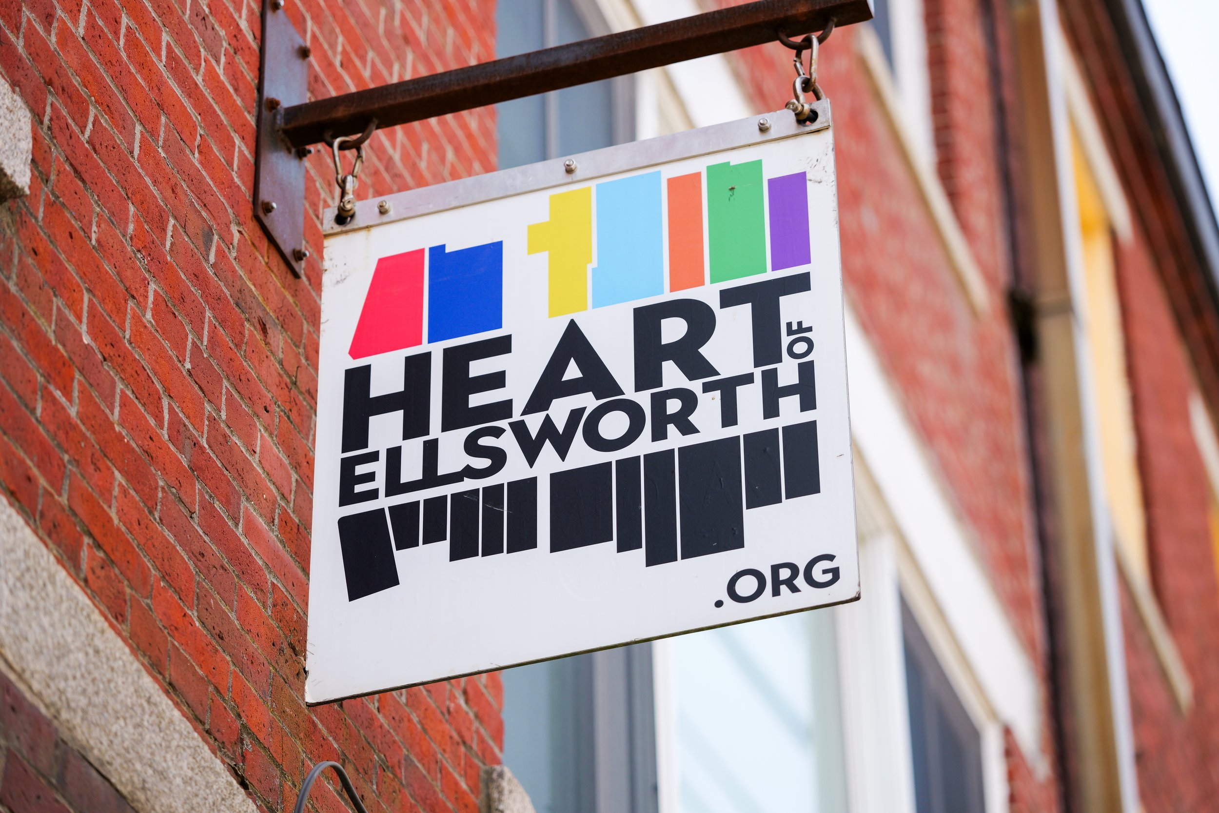A colorful art gallery sign hanging on a red brick building, featuring the text 'ART HEIRS OF HILLSWORTH'. the sign is attached with metal brackets.