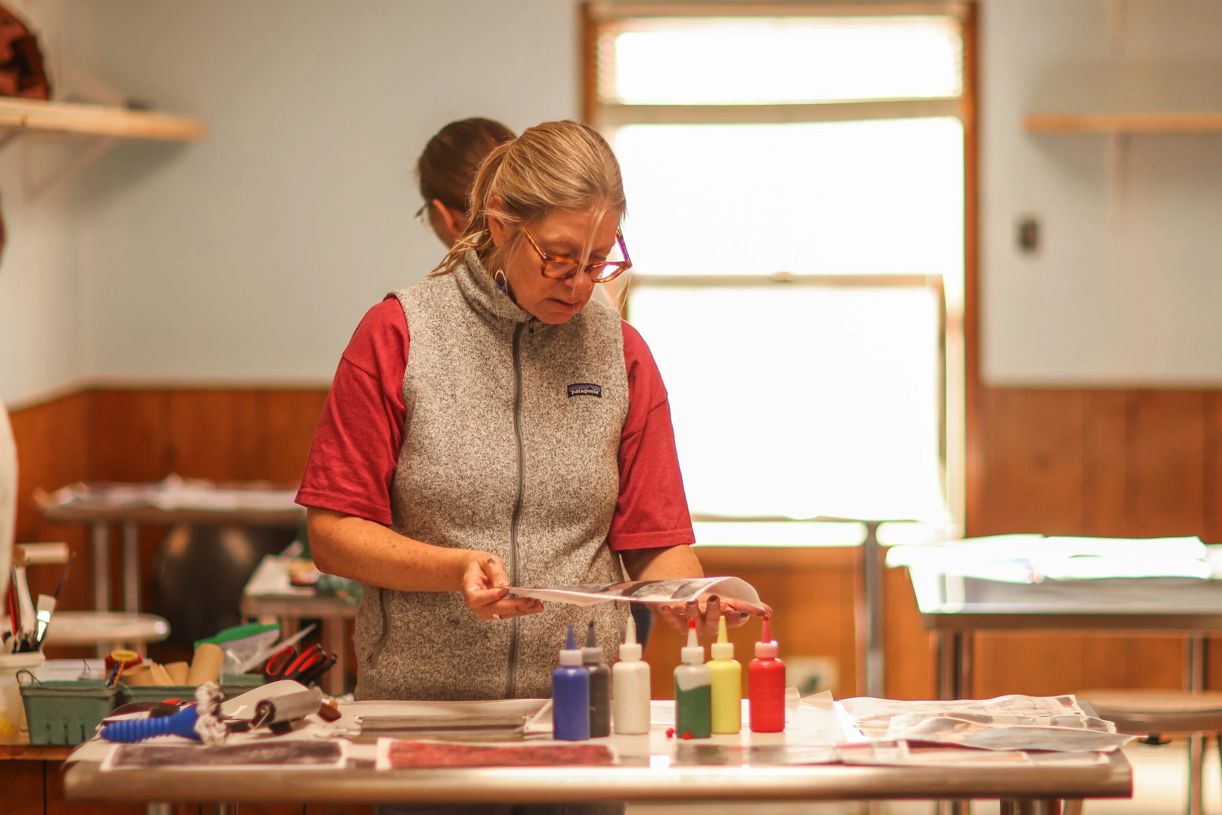 A woman with glasses and blonde hair, wearing a red shirt and beige vest, reading a paper in a room with sunlight coming through a window behind her. There are art supplies and bottles of paint on the table in front of her.