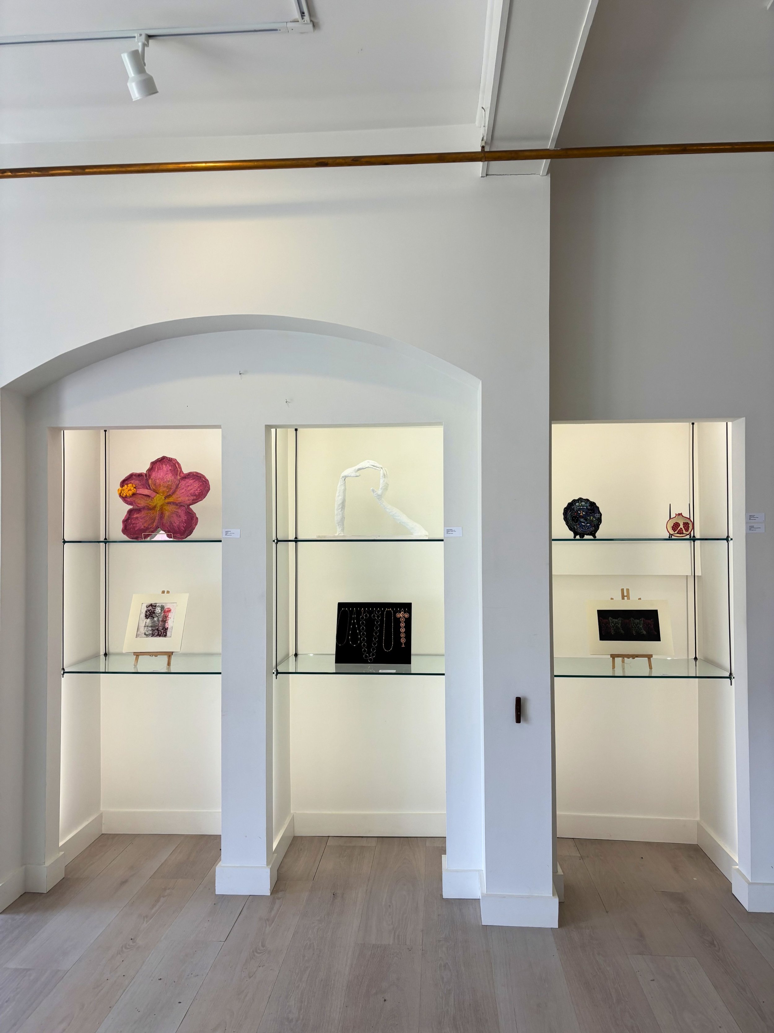 Art gallery display case with various art pieces including a large pink flower, jewelry, and framed artwork, in a room with white walls and light wood floor.
