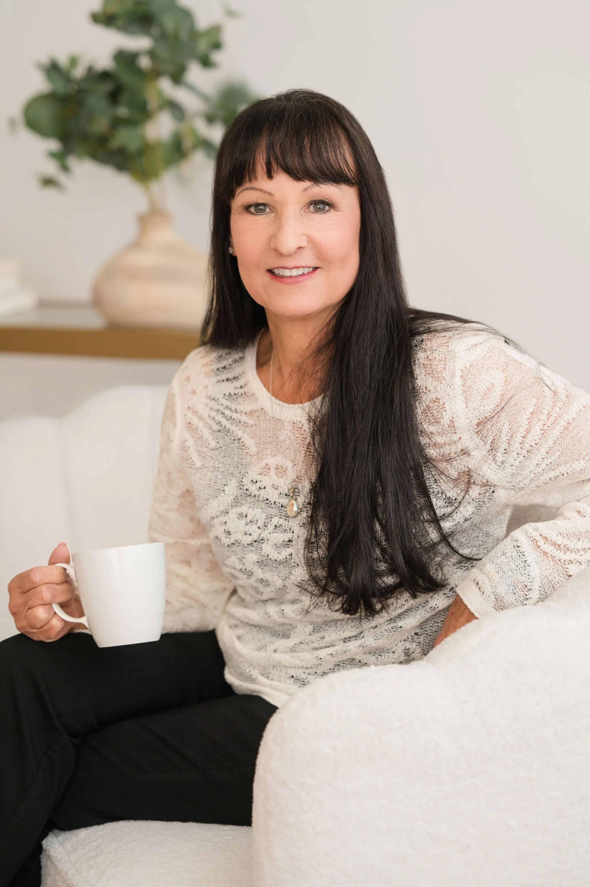 A woman with long dark hair and light skin, smiling and holding a white mug, sitting on a bed with white bedding, in a room with a neutral-colored wall and a plant in a beige vase in the background.