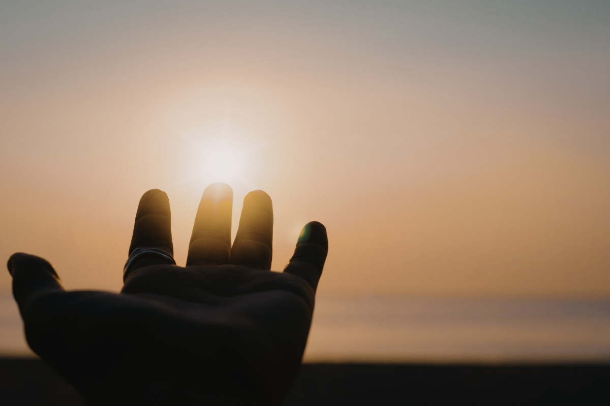 A hand reaching towards the sun with the ocean in the background during sunset