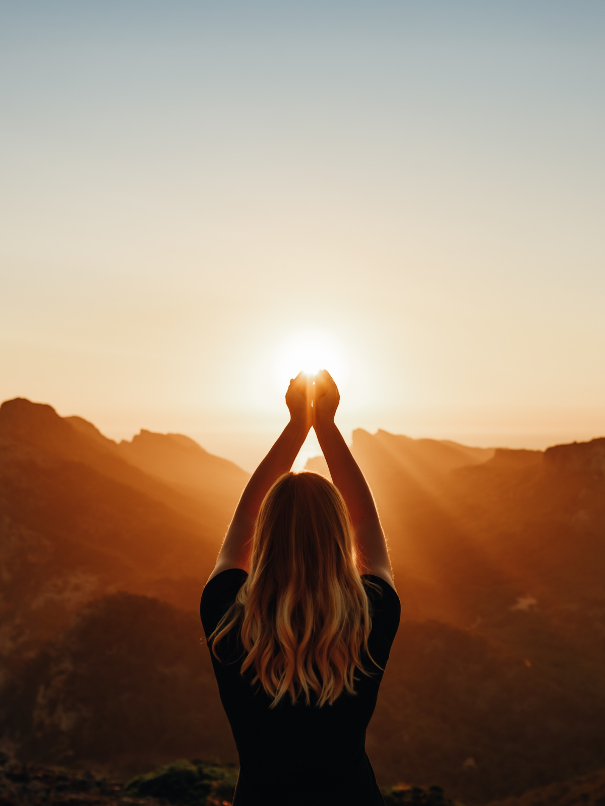 A woman with long blonde hair is standing outdoors at sunset, holding her hands up with palms touching as she faces the sun and mountains in the background.