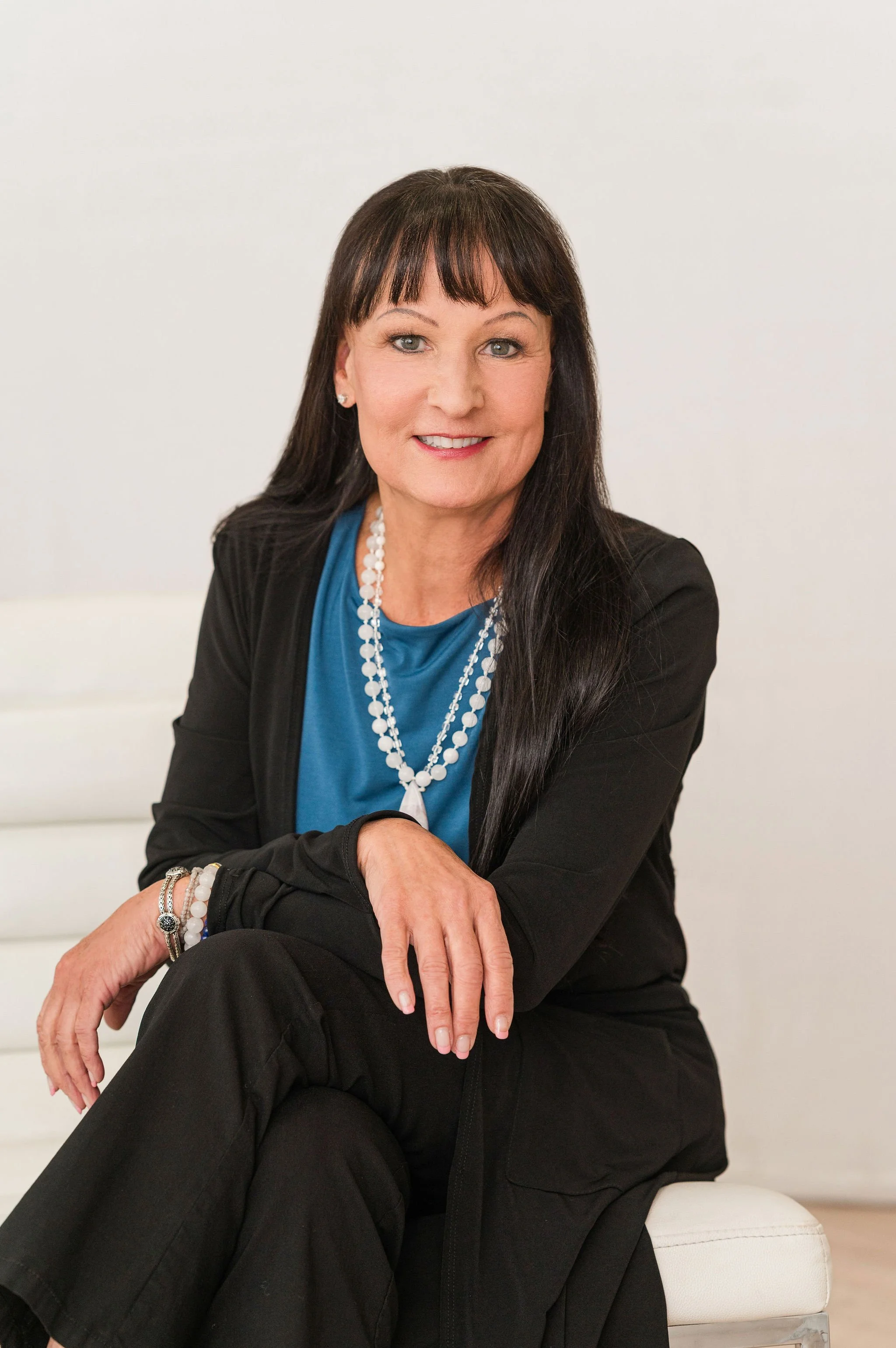A woman with long dark hair and bangs, wearing a blue blouse, a black blazer, and jewelry, sitting on a white couch against a plain white wall.