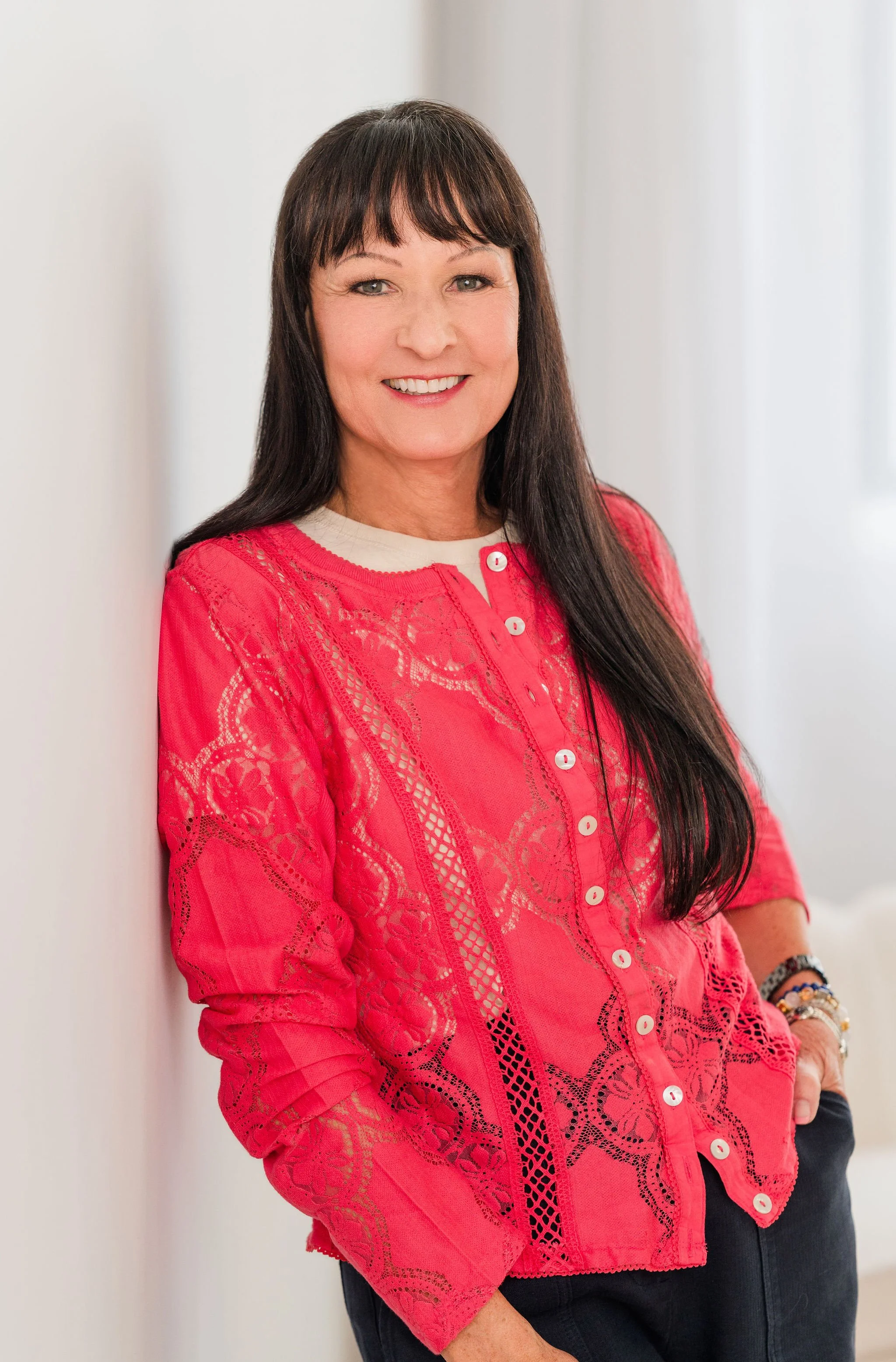 A woman with long dark hair and bangs, wearing a bright pink lace blouse with white buttons, standing against a white wall, smiling, with bracelets on her right wrist.