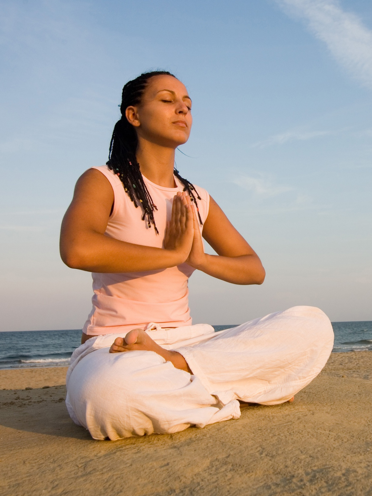 Woman practicing yoga outdoors on the beach, sitting cross-legged in a meditative pose with hands pressed together in prayer position and eyes closed.