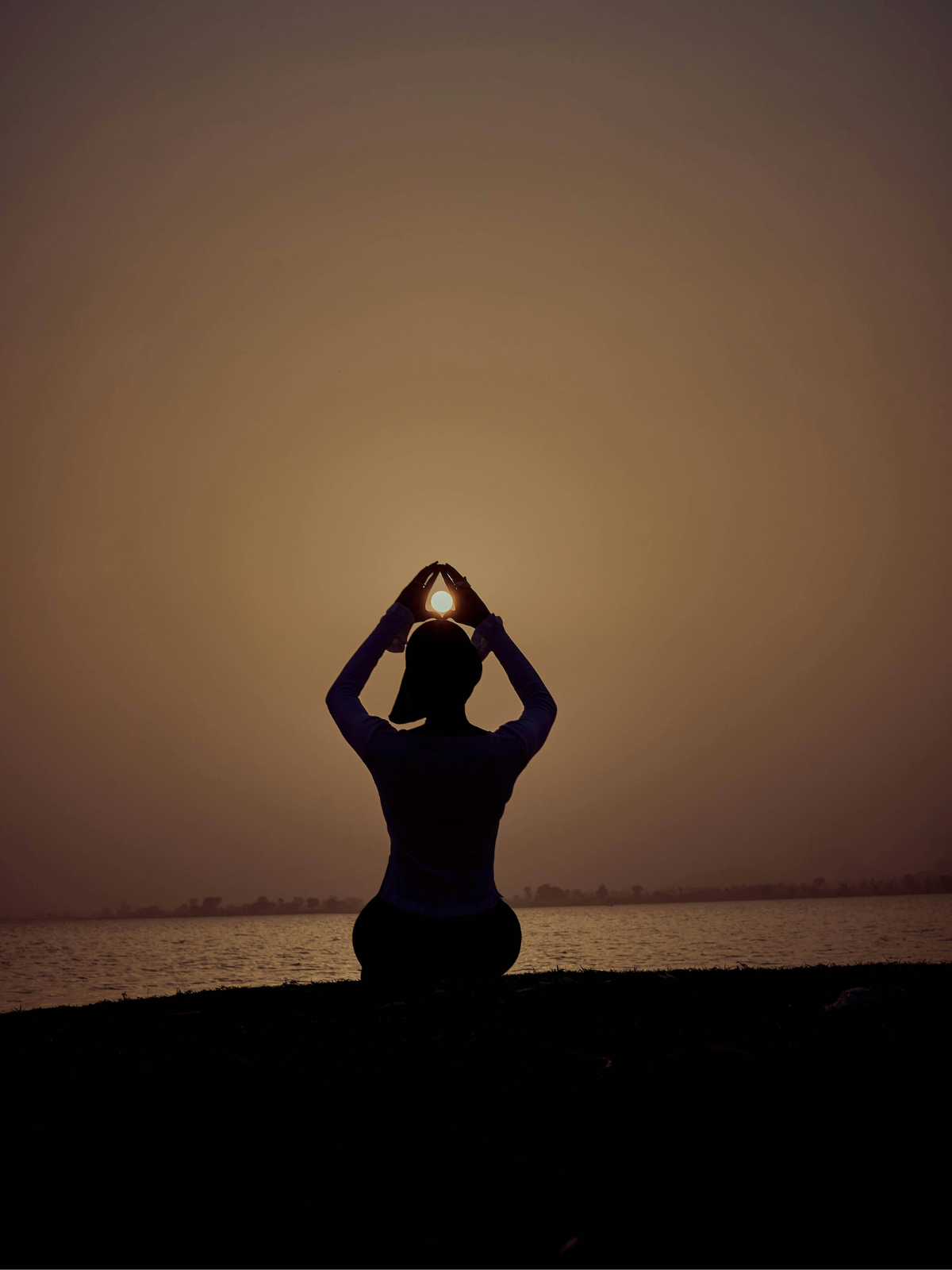 Silhouette of a person sitting cross-legged outdoors during sunset, holding hands above head holding a glowing light.