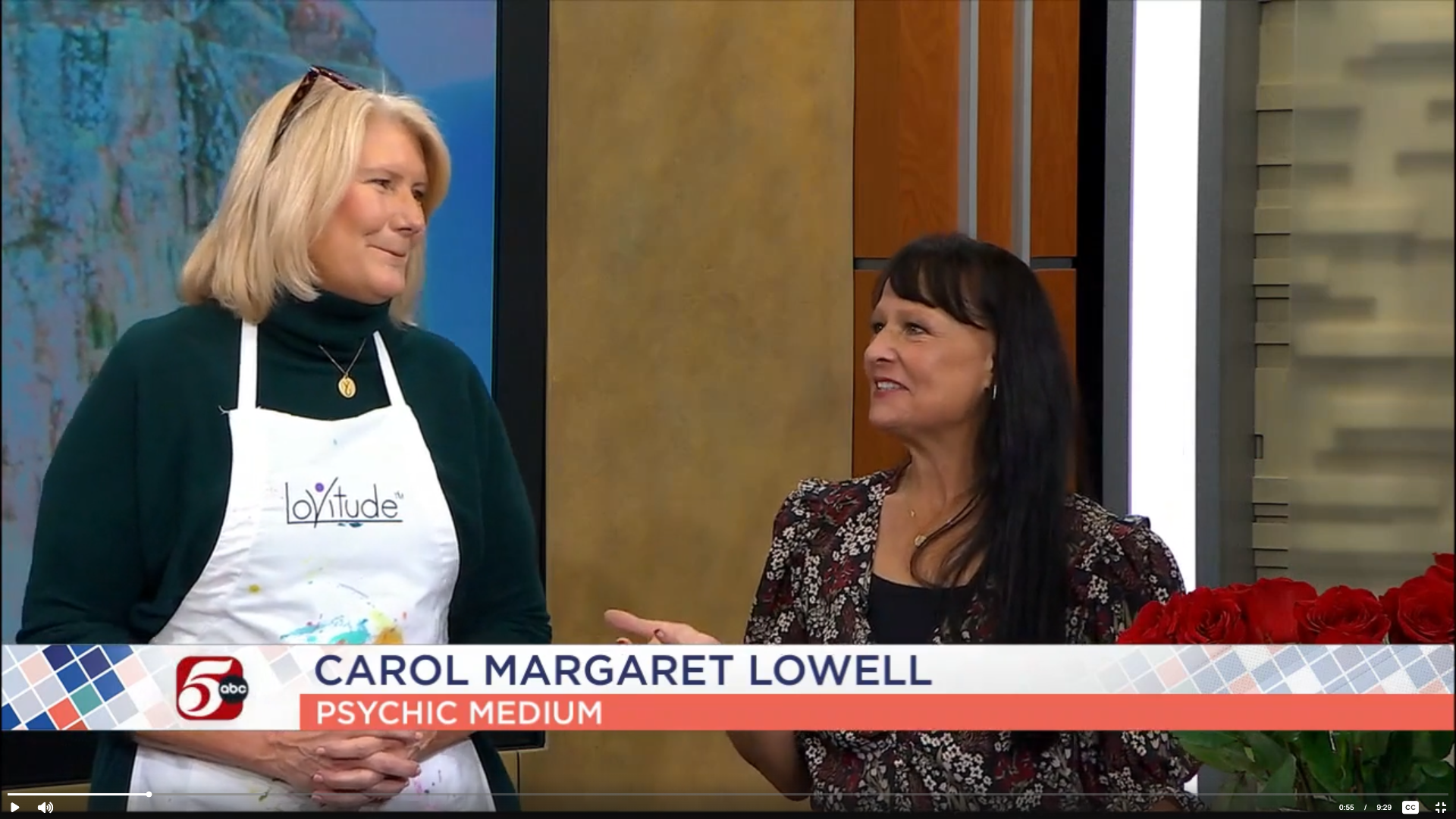 Two women are talking on a TV set with a news banner at the bottom. One woman, with blonde hair, is wearing a black green turtleneck and a white apron. The other woman, with dark hair, is wearing a floral blouse and is smiling. There is a large bouquet of red roses on a table in front of them.