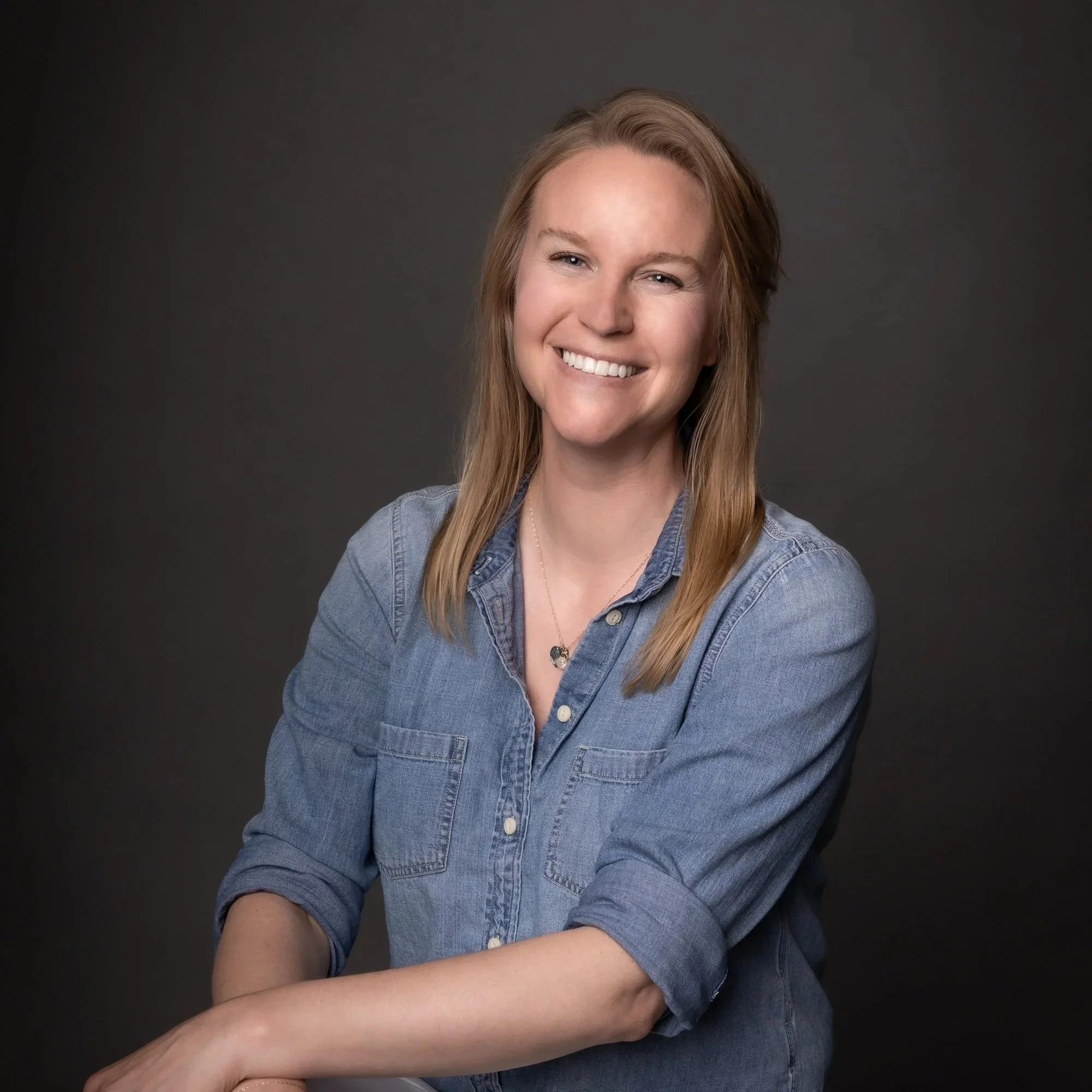 A woman with long blonde hair, smiling, wearing a denim shirt, sitting with her arms crossed against a dark gray background.