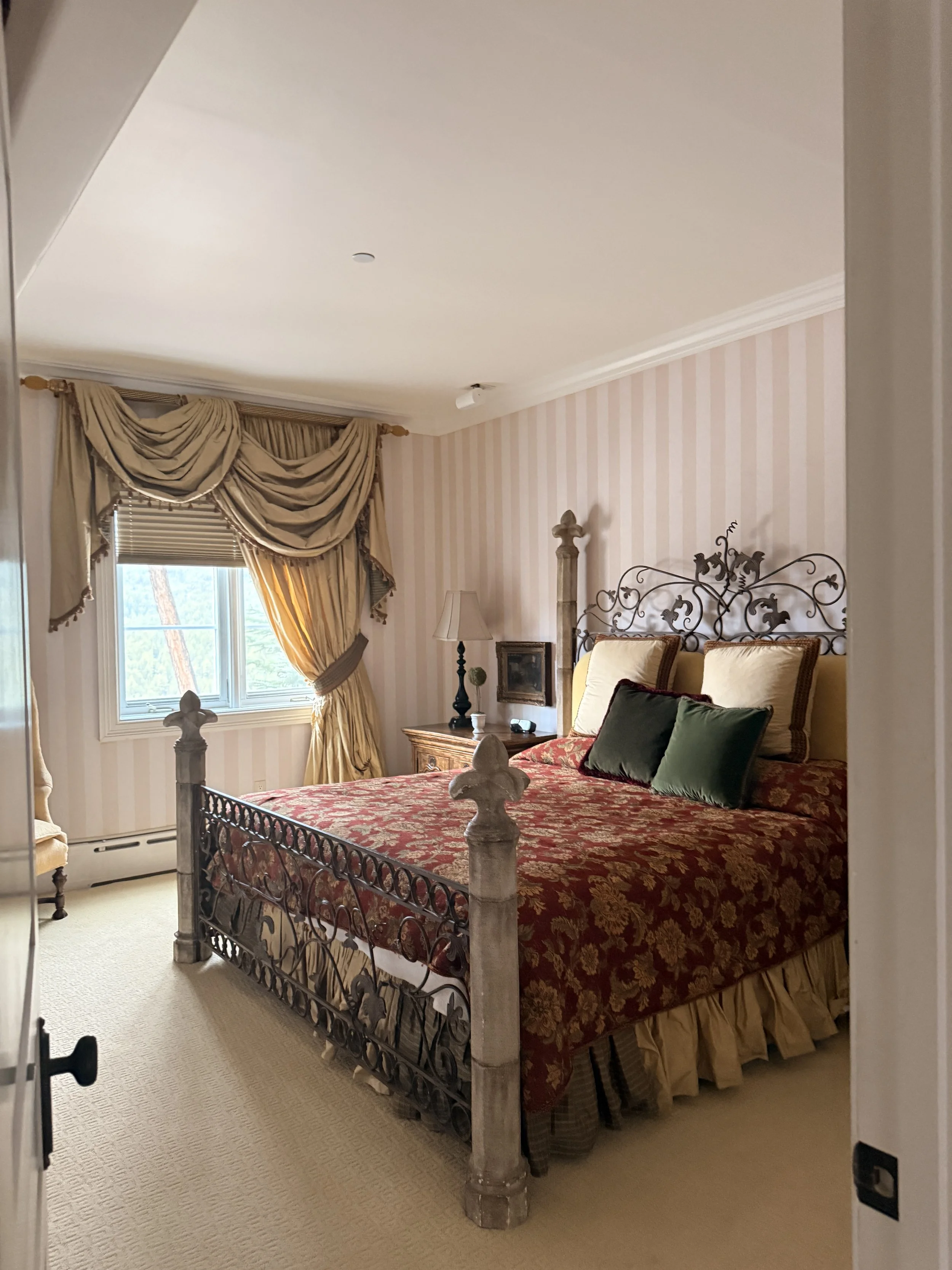 A bedroom with a four-poster bed, ornate headboard, yellow and red patterned bedding, and multiple pillows. There are window curtains with gold and beige drapes, a nightstand with a lamp, and a window showing trees outside.