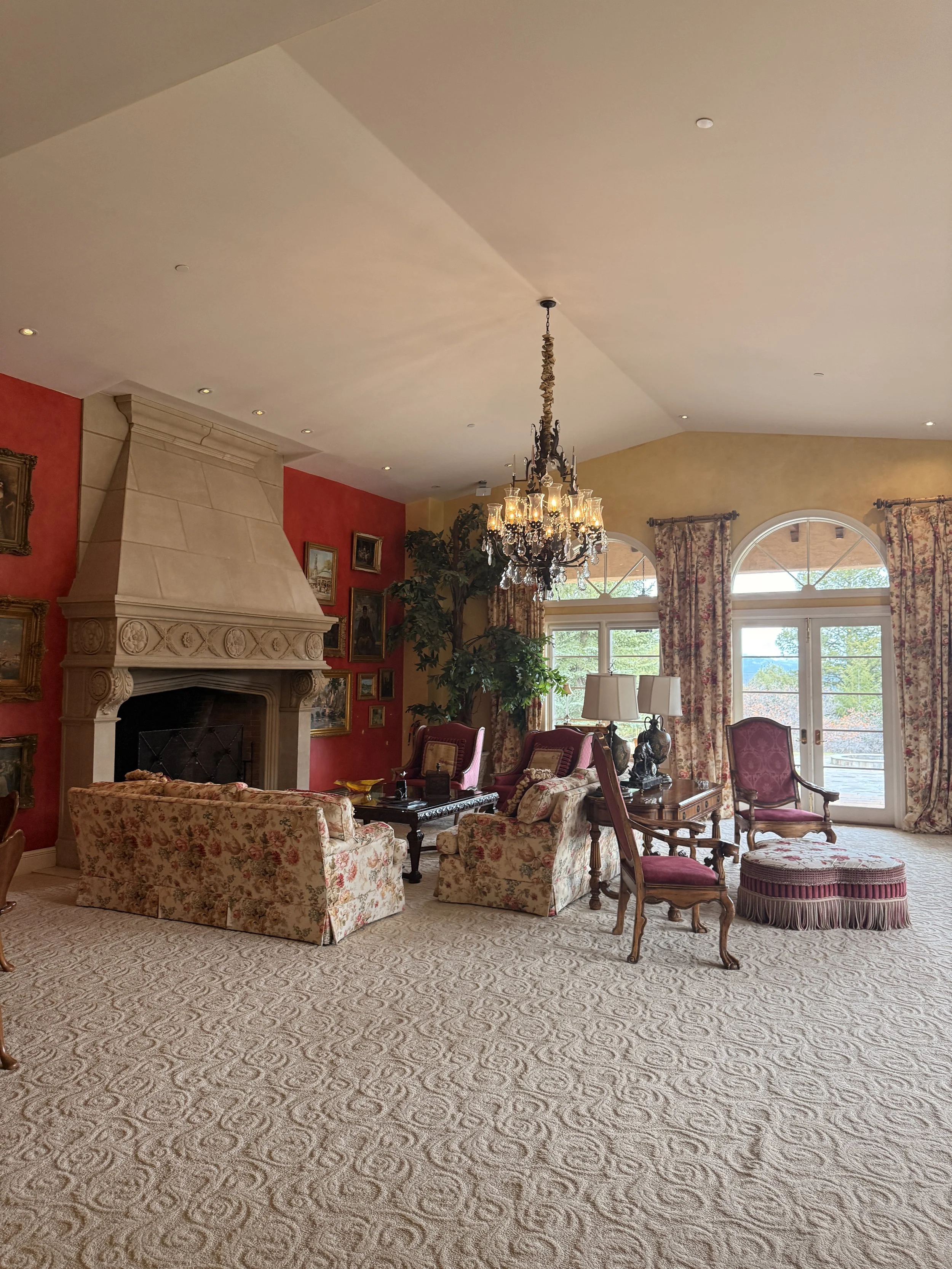 A spacious living room with ornate furniture, a large fireplace, chandeliers, and large windows with floral curtains, decorated in warm tones.