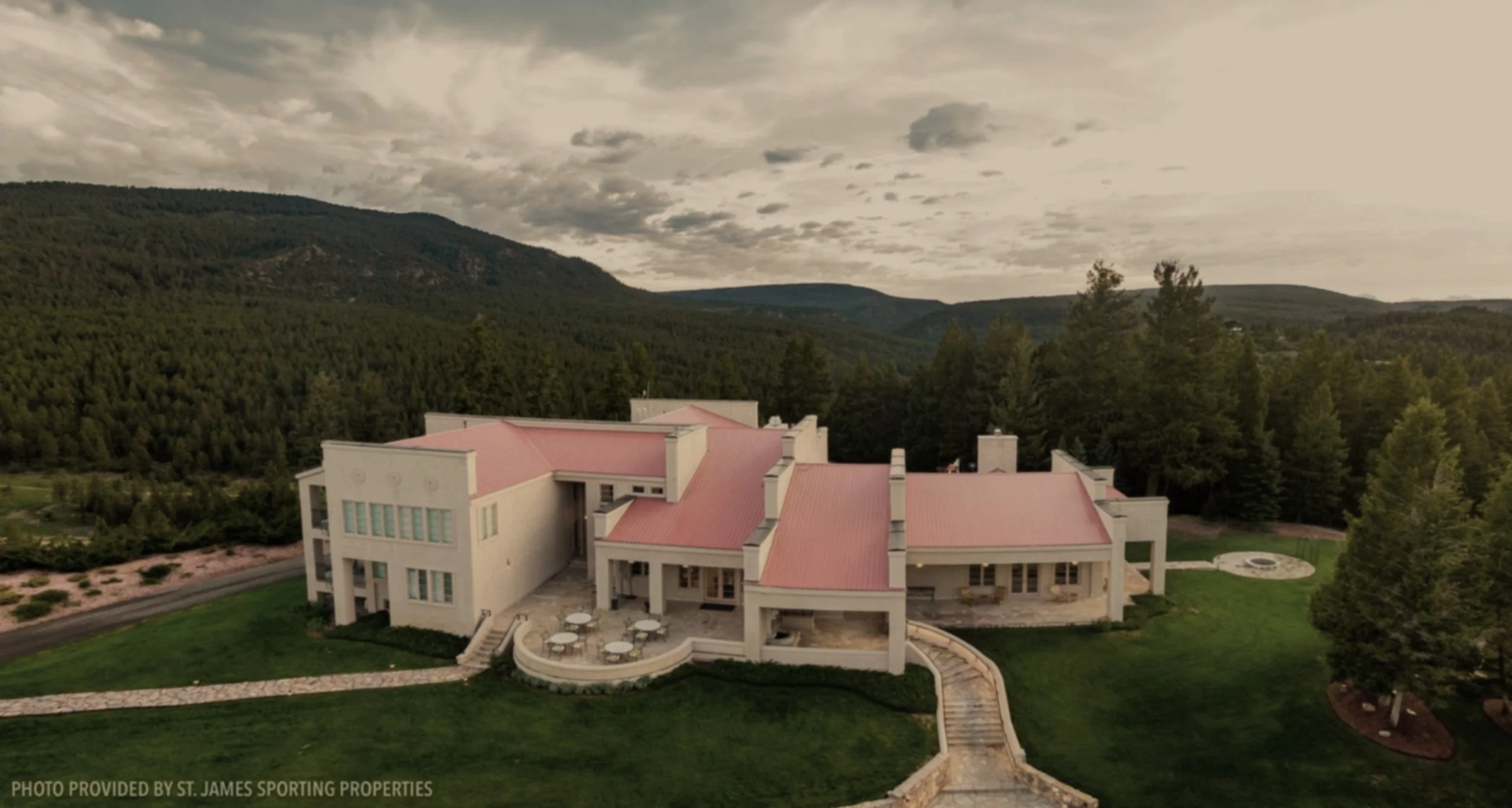 An aerial view of a large, modern house with white walls and pink roofs, surrounded by green lawns, trees, and a mountainous landscape under a cloudy sky.