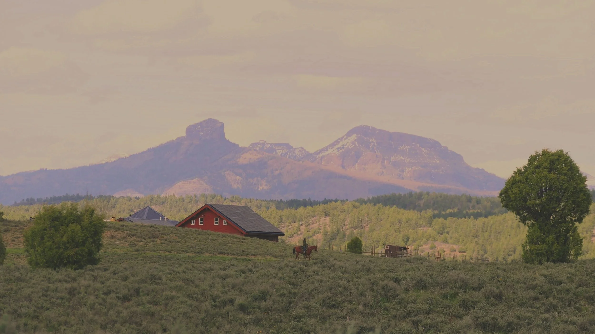 A landscape with mountains in the background, a red house and green trees in the foreground, and a person riding a horse.