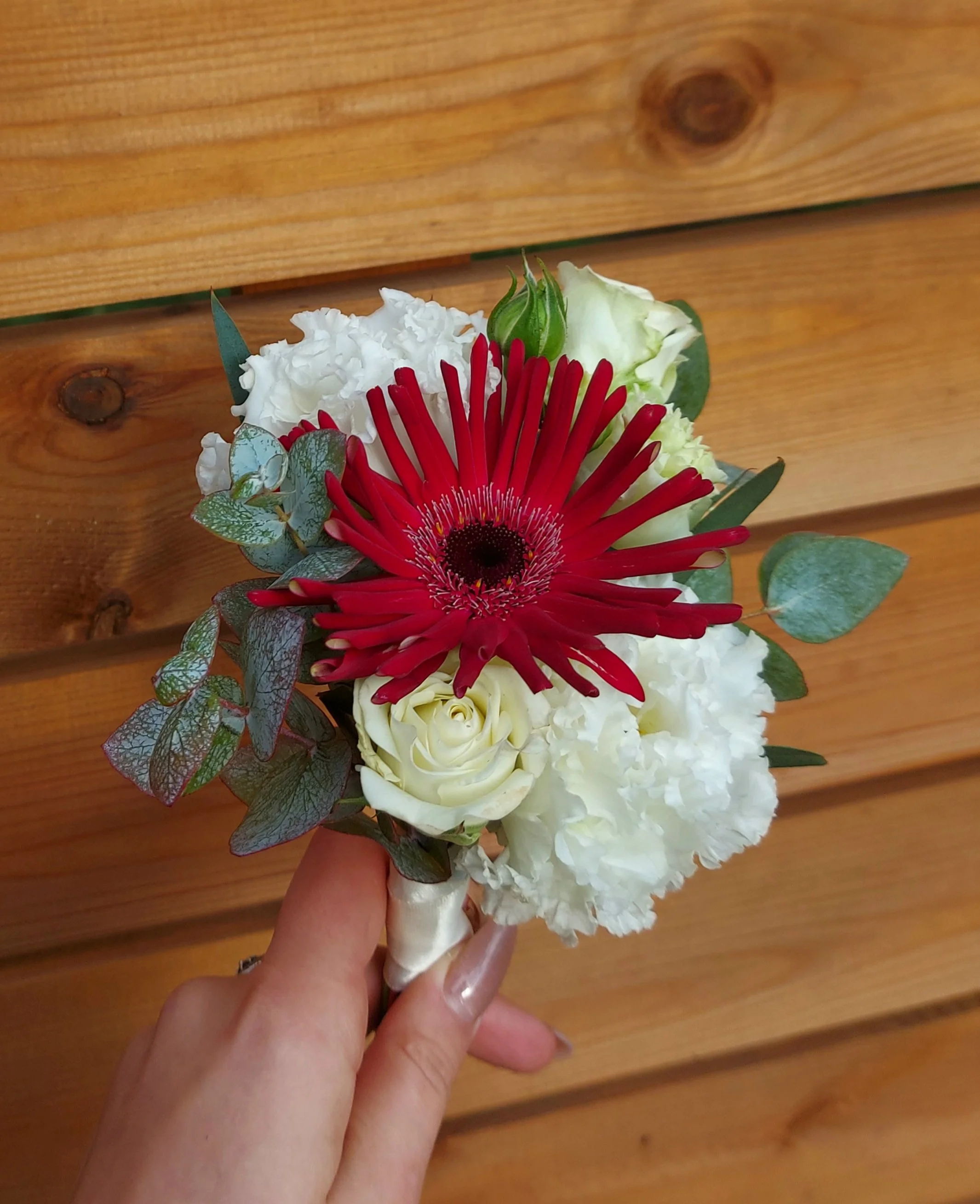 A hand holding a small bouquet of white roses, red gerbera daisy, eucalyptus leaves, and other small flowers against a wooden background.