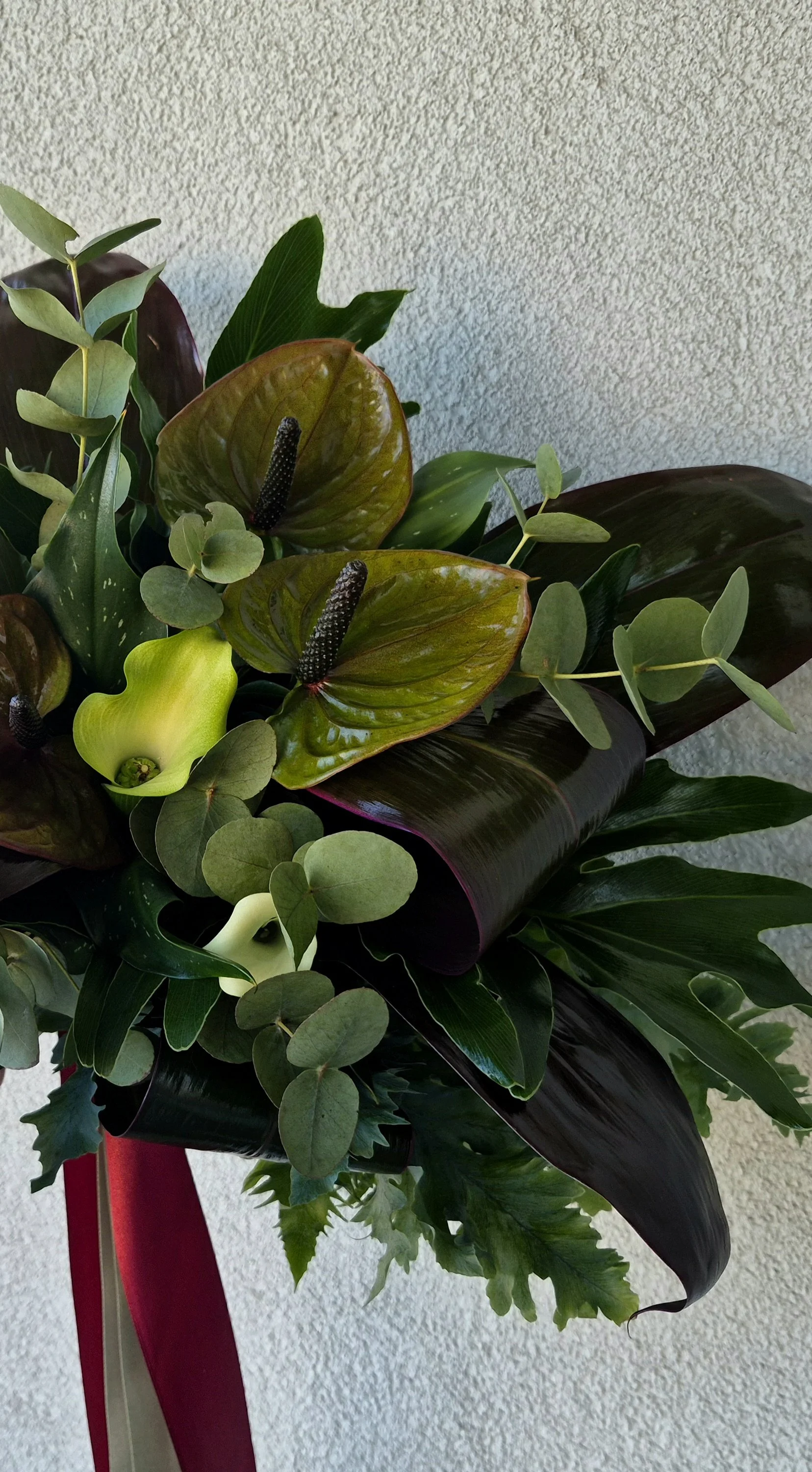 A bouquet of various green and dark-colored leaves with two light green calla lilies and two brownish anthurium flowers with black spadixes, set against a textured beige wall.