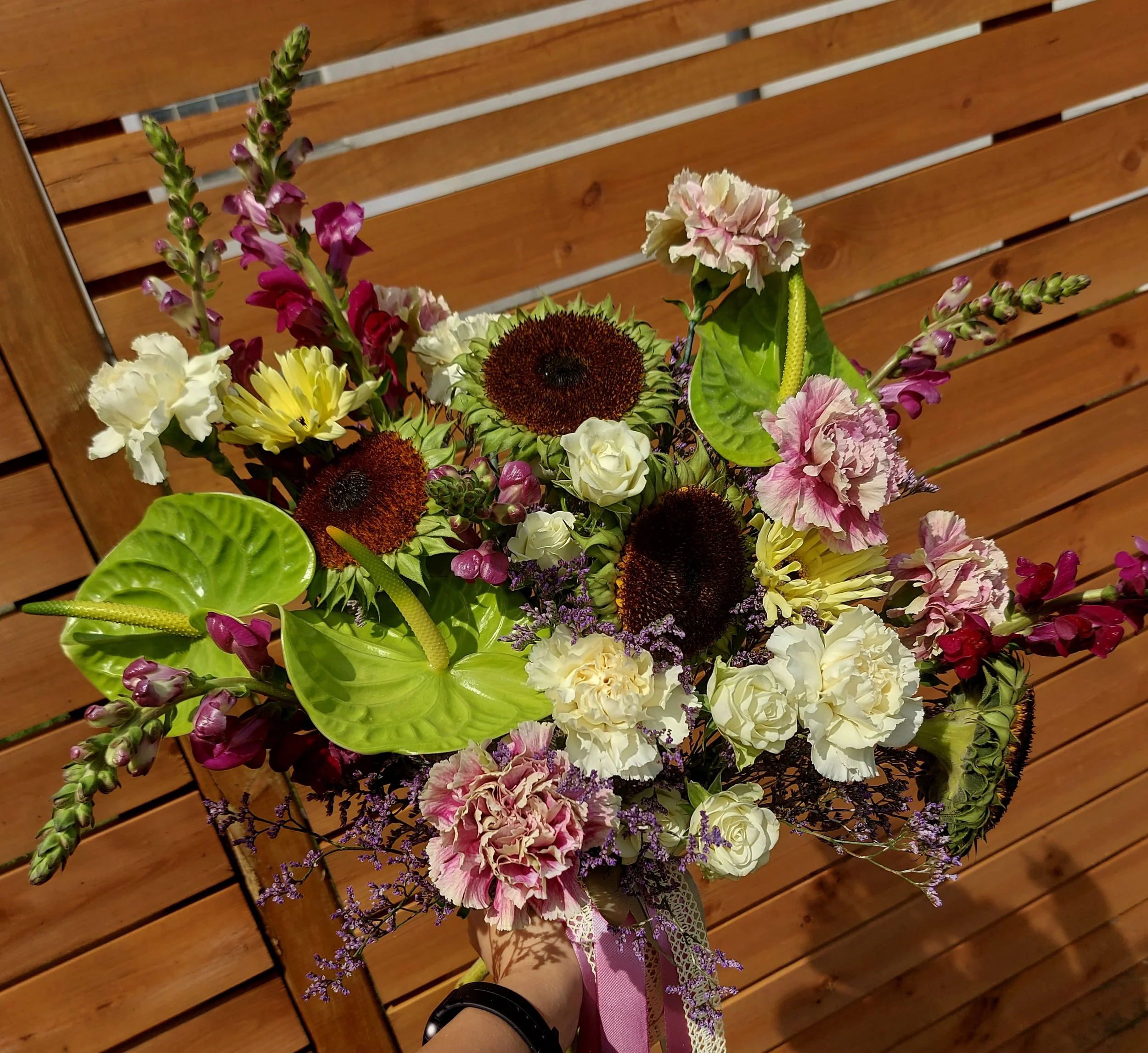 A colorful bouquet of flowers including sunflowers, roses, carnations, and lavender, held by a person wearing a pink ribbon with lace, against a wooden slat background.