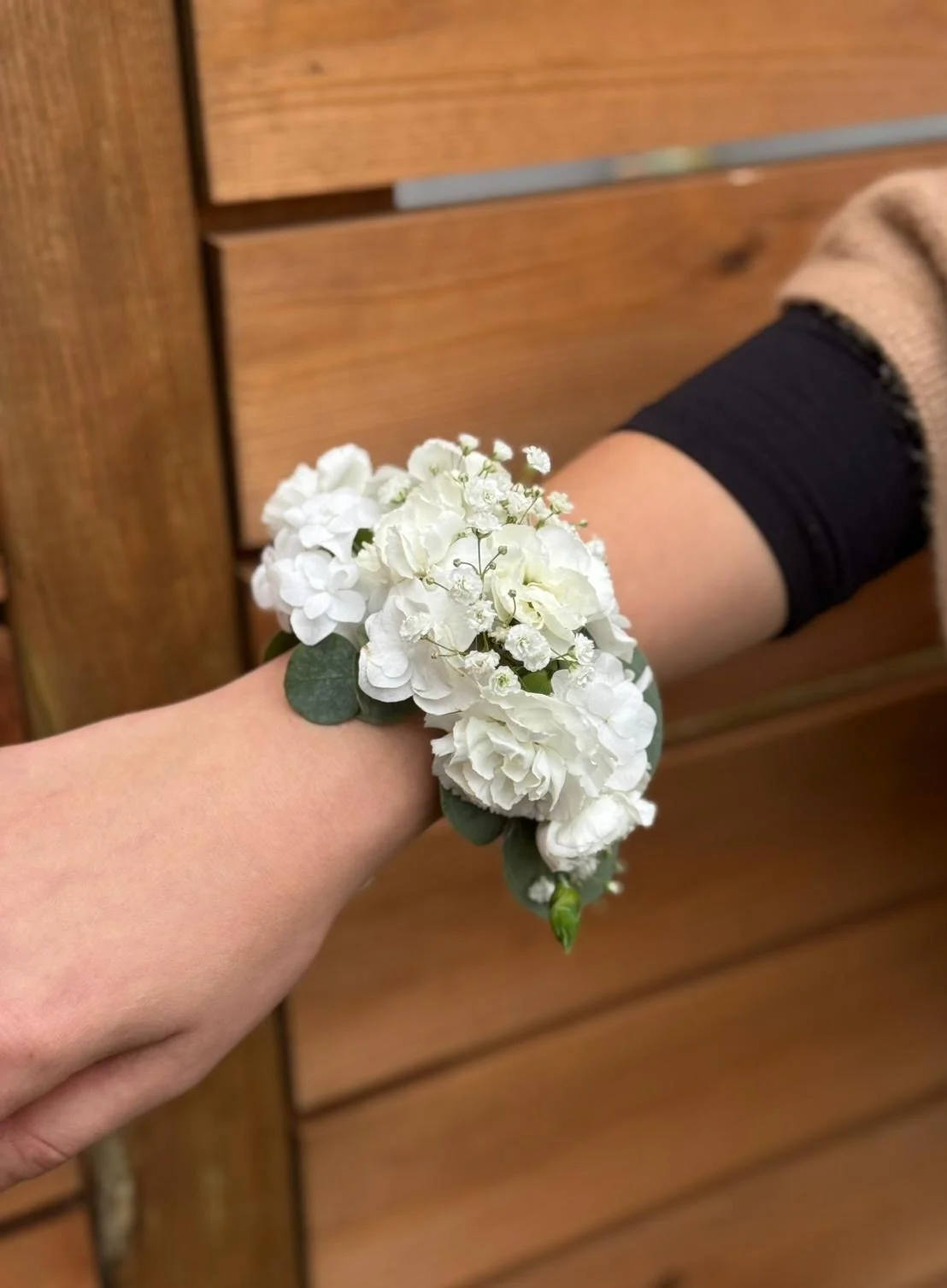 Person wearing a floral corsage on their wrist, holding hands with another person, against a wooden background.