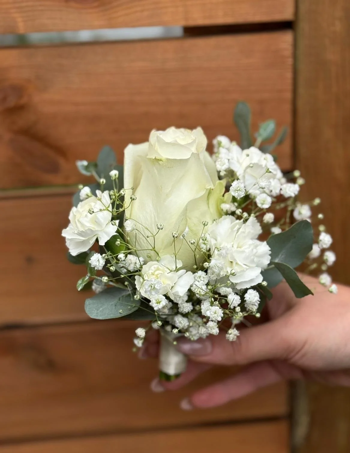 Hand holding a small bouquet of white roses, baby's breath, and eucalyptus leaves against a wooden background.