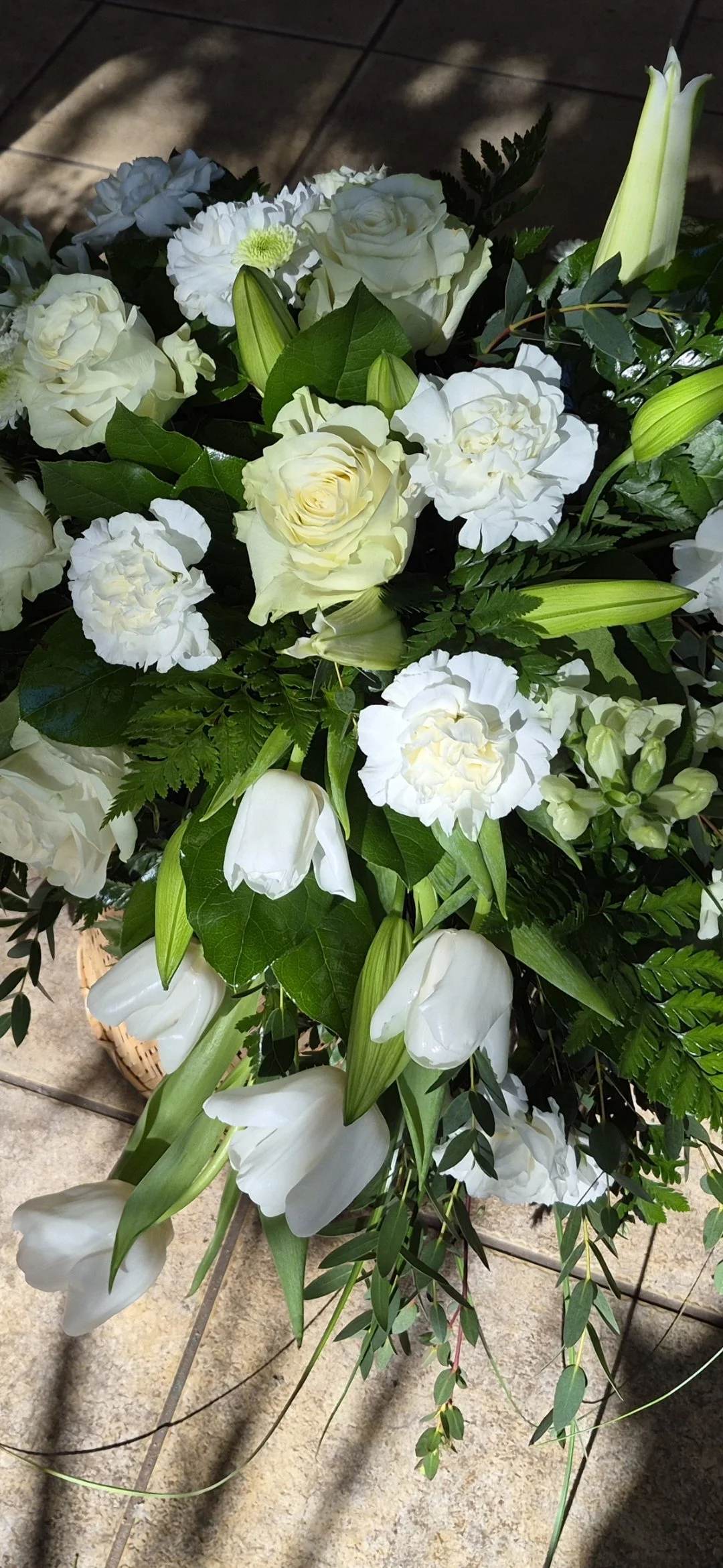 A bouquet of white flowers including roses, lilies, and carnations with green leaves on a tiled floor.