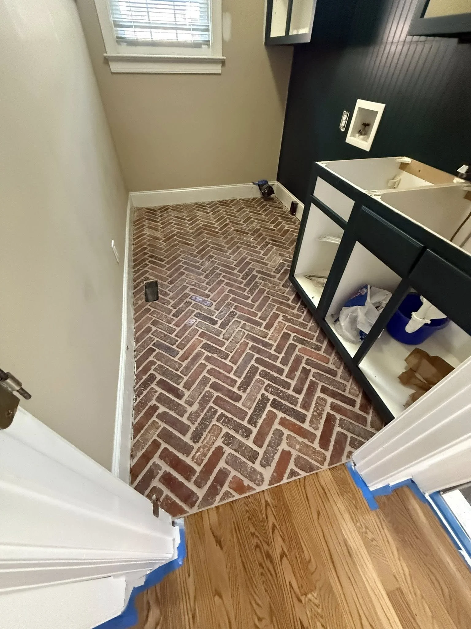 Photo of a small room or corner with a brick-patterned floor, an electrical outlet, a window with blinds, and a black wall with kitchen cabinets. Part of the floor is covered with blue painter's tape near the edge of a different hardwood floor.