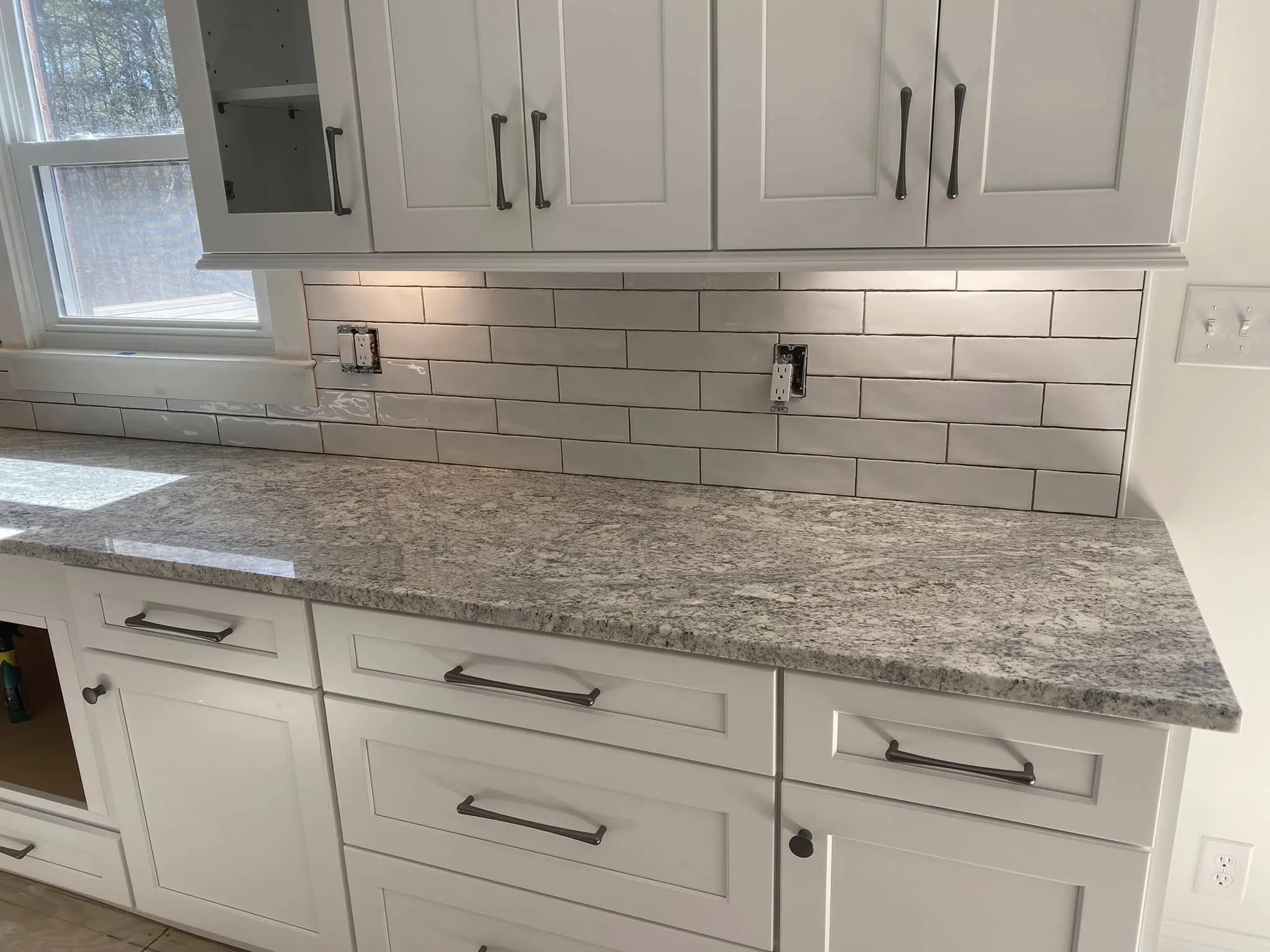 Kitchen with white cabinetry, a granite countertop, a tiled backsplash, and a window with natural light. There are electrical outlets and outlets for appliances without covers.