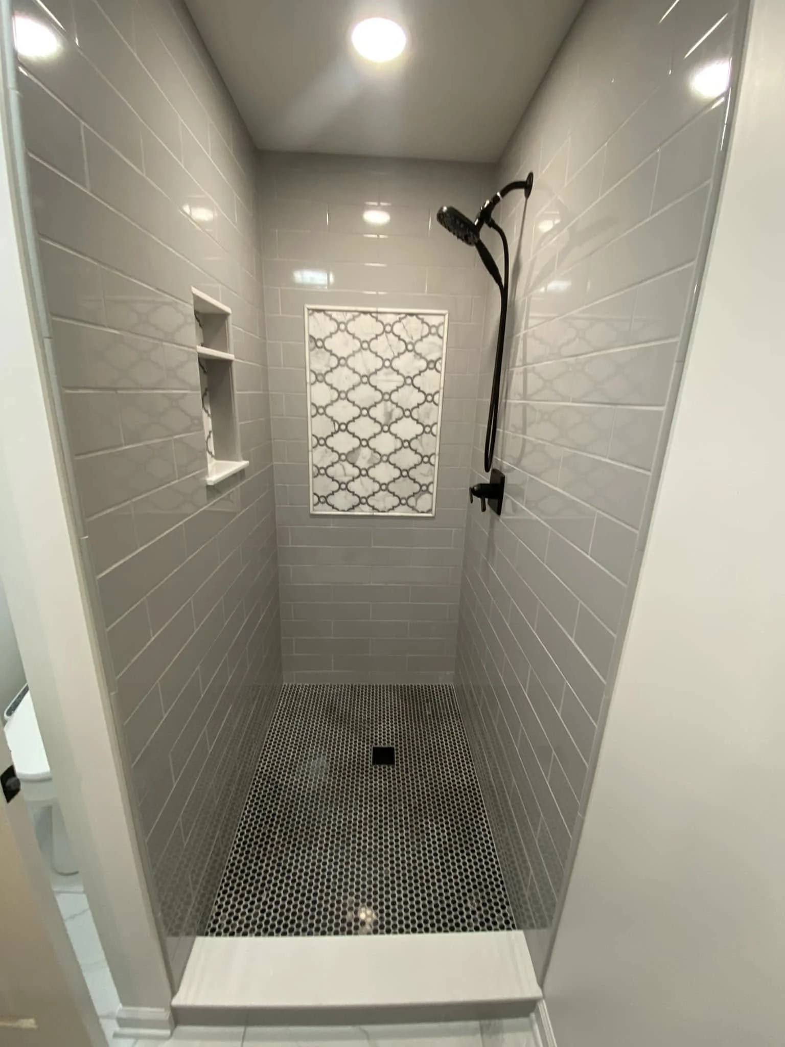 Modern shower with gray tiled walls, a patterned inset, a black showerhead, and a black handheld sprayer, with a black drain at the floor.