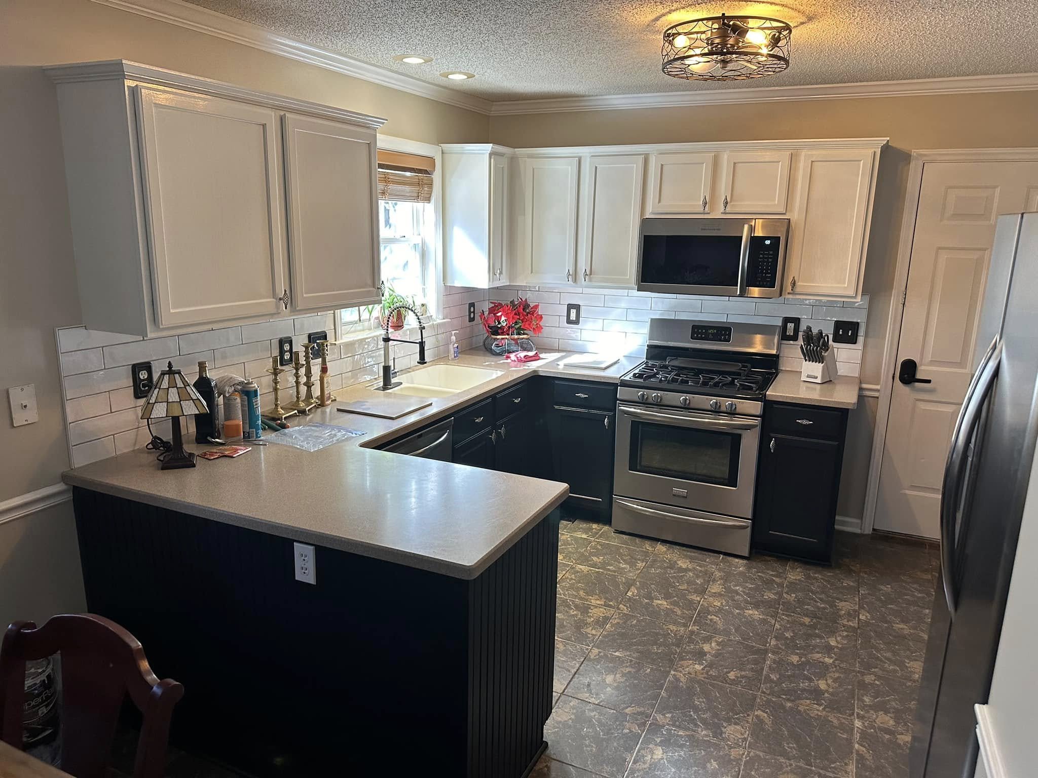 Kitchen with white upper cabinets, dark lower cabinets, stainless steel oven and microwave, beige countertop, tiled backsplash, window with blinds, poinsettia plant, and various small items on countertop.