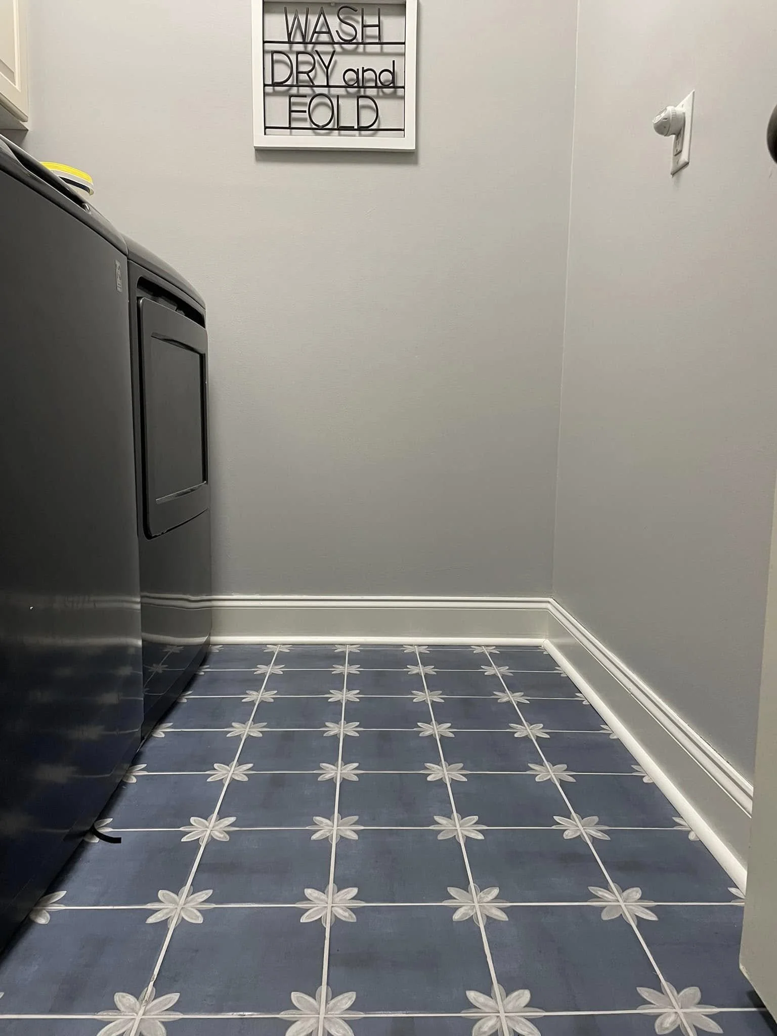 A laundry room with a washing machine on the left, a white wall with a sign that says 'WASH DRY and FOLD' above, gray walls, a power outlet on the right, and blue and white floral patterned floor tiles.