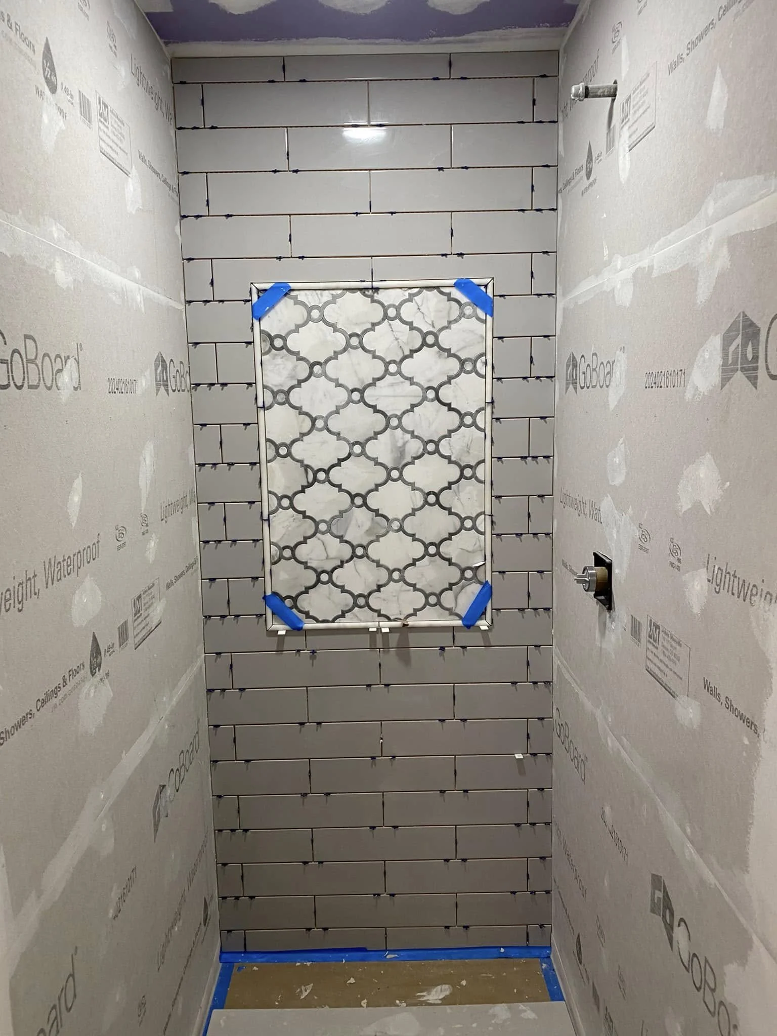 A small bathroom under construction with wall tiles and a decorative tile panel at the back wall, surrounded by drywall with protective covering.