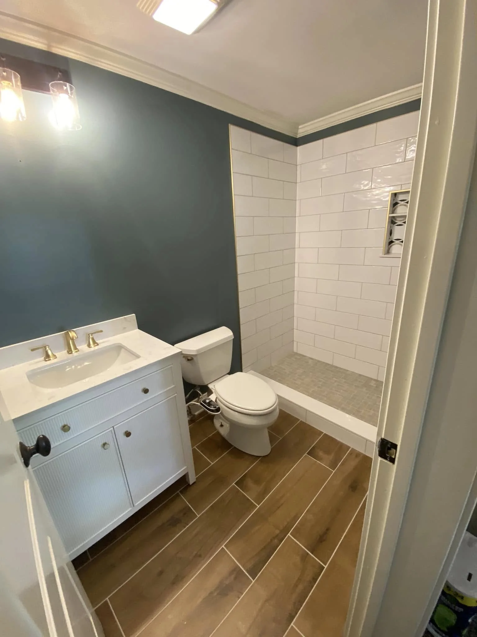 A small bathroom with wood-look tile flooring, a white vanity with gold fixtures, a toilet, and a walk-in shower with white subway tile walls and a small built-in niche.