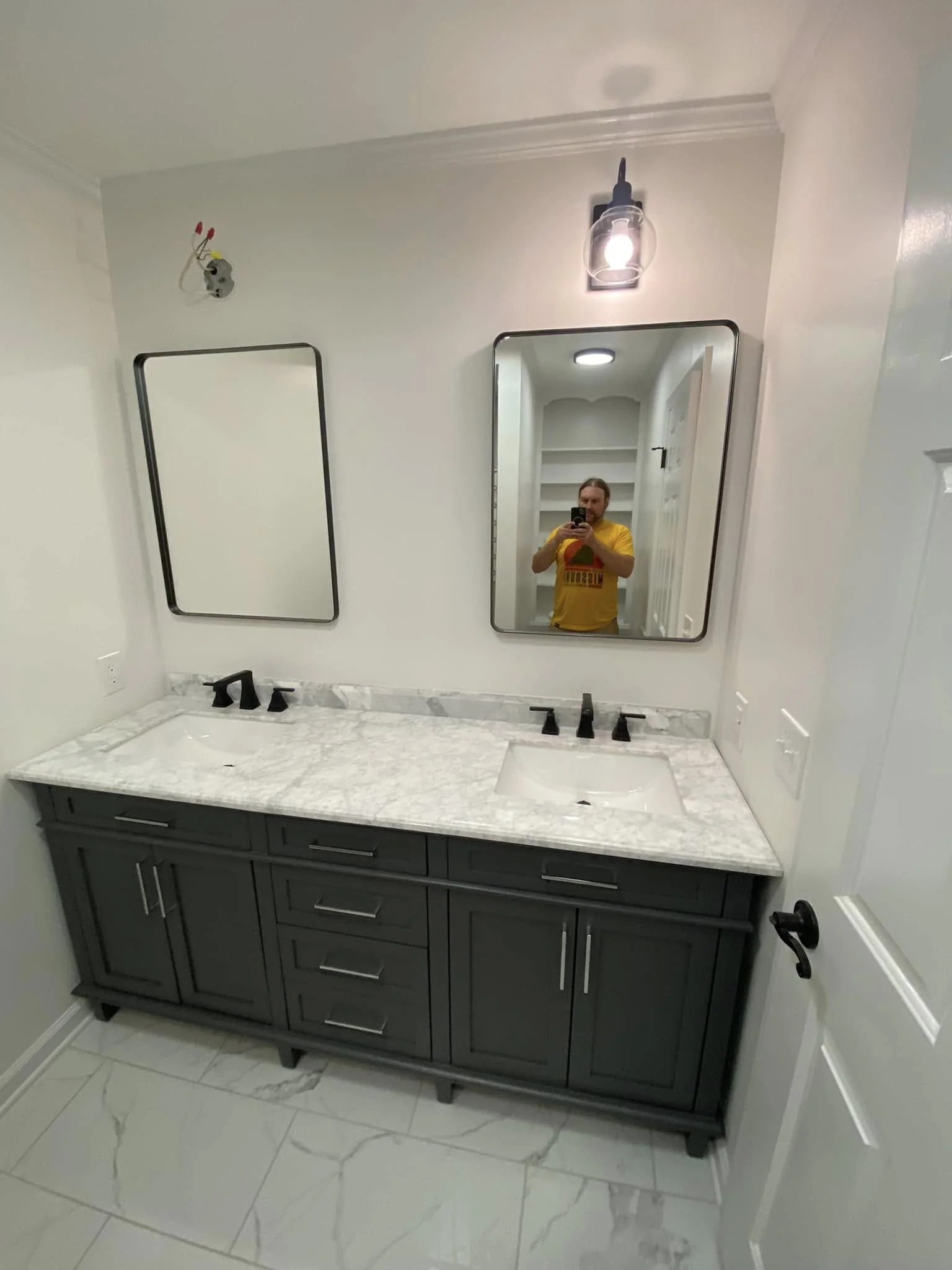 Bathroom vanity with dark gray cabinet, white marble countertop, and two mirrors, with a person taking a photo in the reflection.