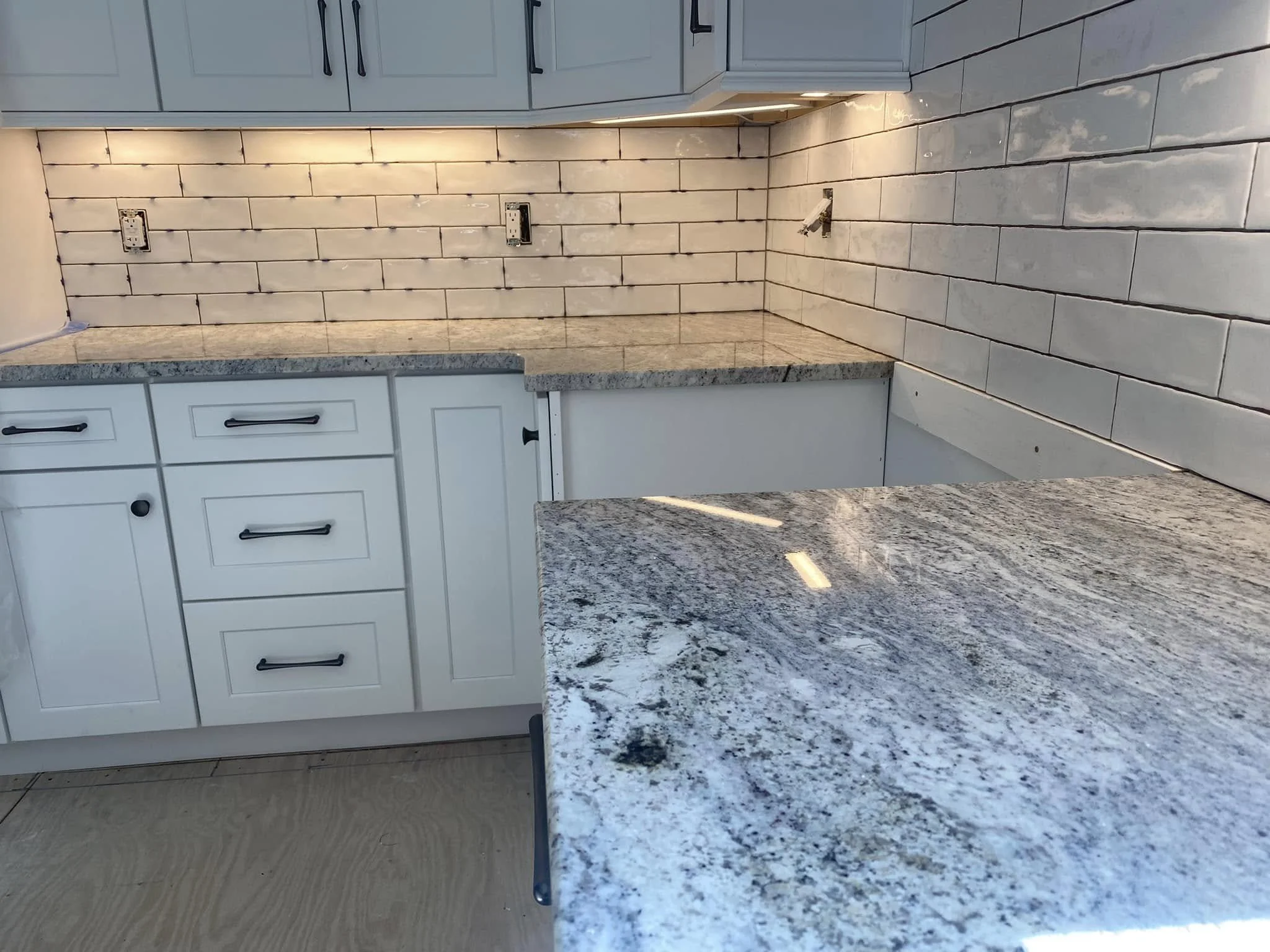 Kitchen with light-colored cabinets, an under-cabinet light, and a granite countertop. The wall is tiled with bricks, but some outlets are uncovered, and there is a hole where an appliance might be installed.