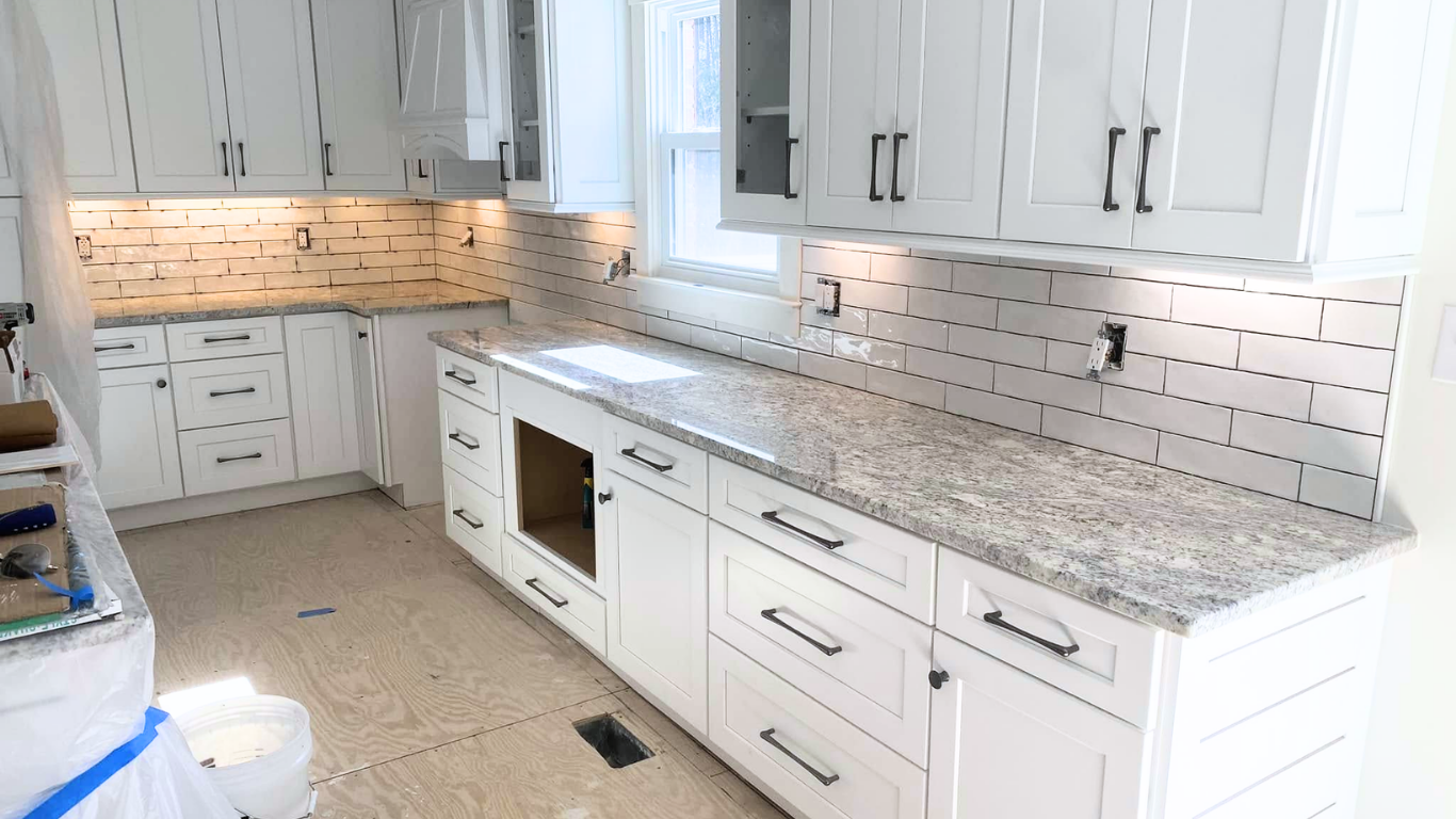 White kitchen cabinets with black handles, granite countertop, unfinished floor, and a tile backsplash.