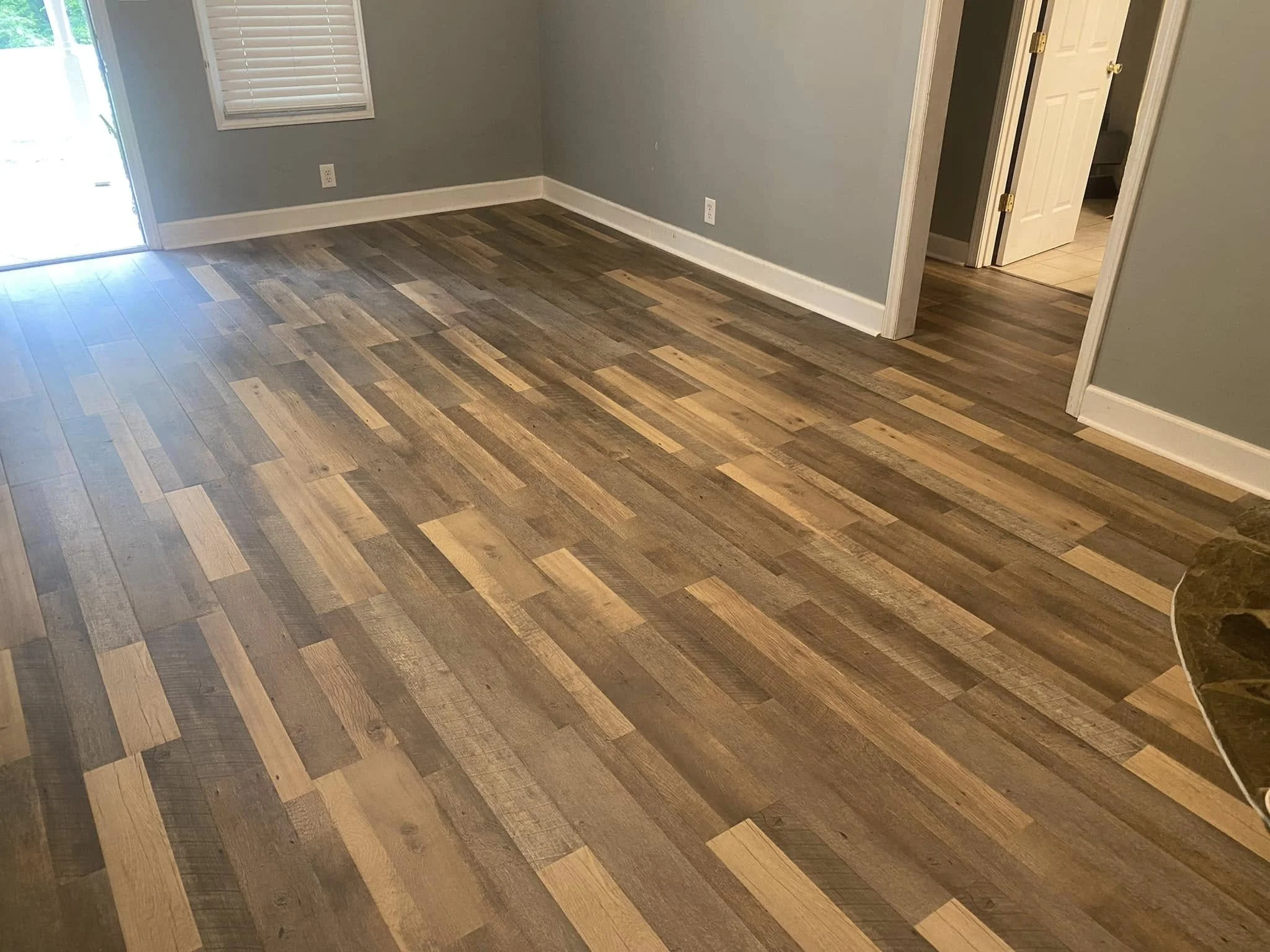 Empty room with wooden flooring, light gray walls, a window with blinds, and an open door leading to another room with tiled floor.