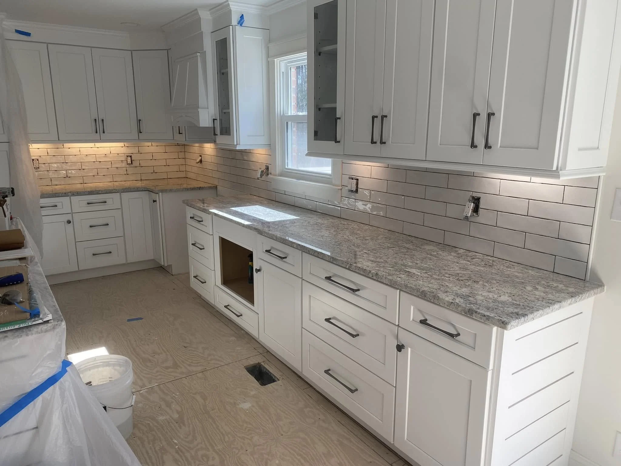 A kitchen under renovation with white cabinets, granite countertops, and a brick-style backsplash. The floor is unfinished, showing plywood subfloor. There are no appliances installed yet, and protective plastic covers are on some surfaces.