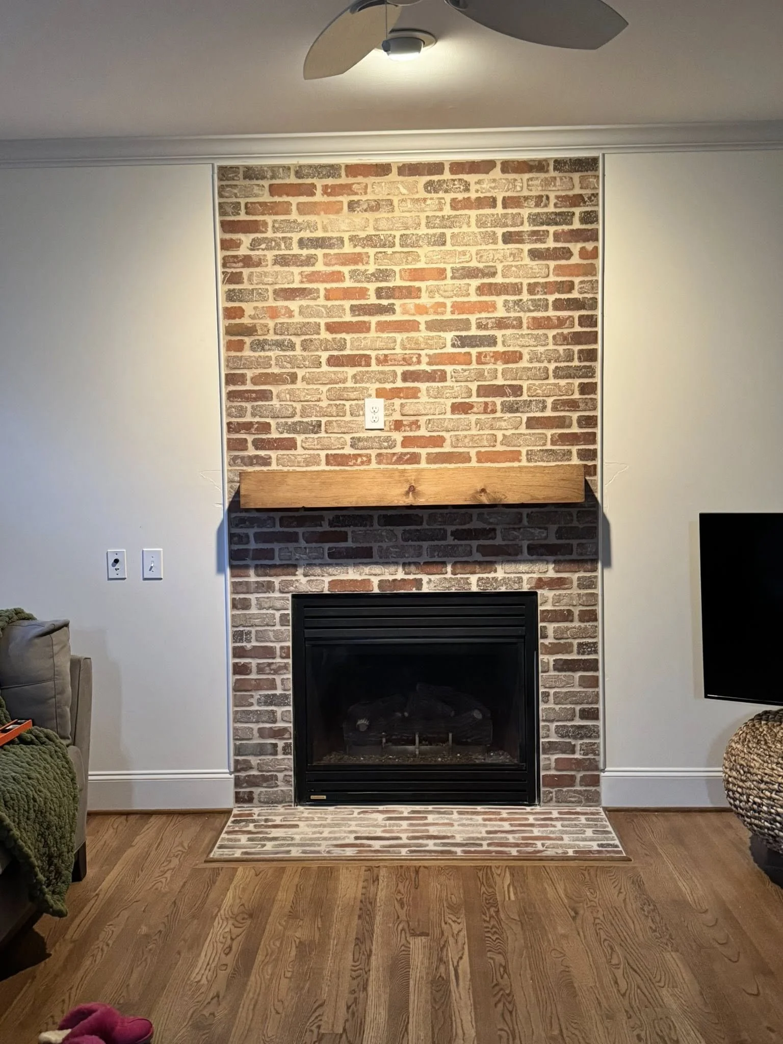 Living room with brick fireplace, wooden mantel, rug, hardwood floor, and partially visible furniture on sides.