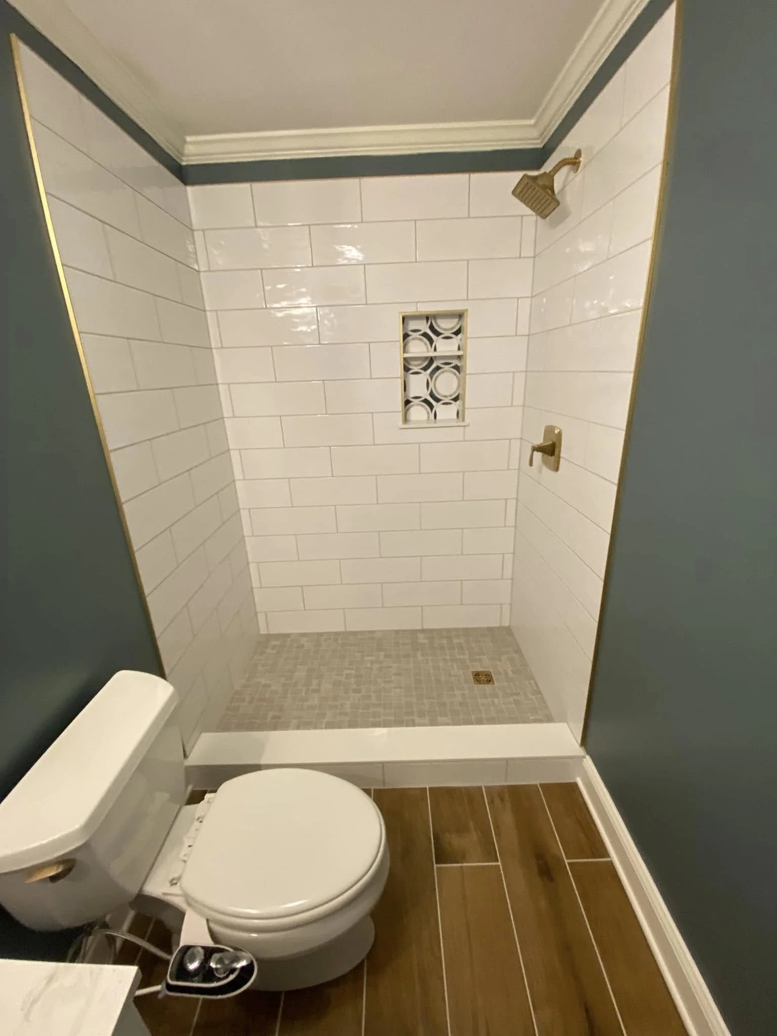 A bathroom with a white toilet, a gold shower head, a gold faucet, cream tile walls, wood flooring, a small window with decorative grille, and teal walls.