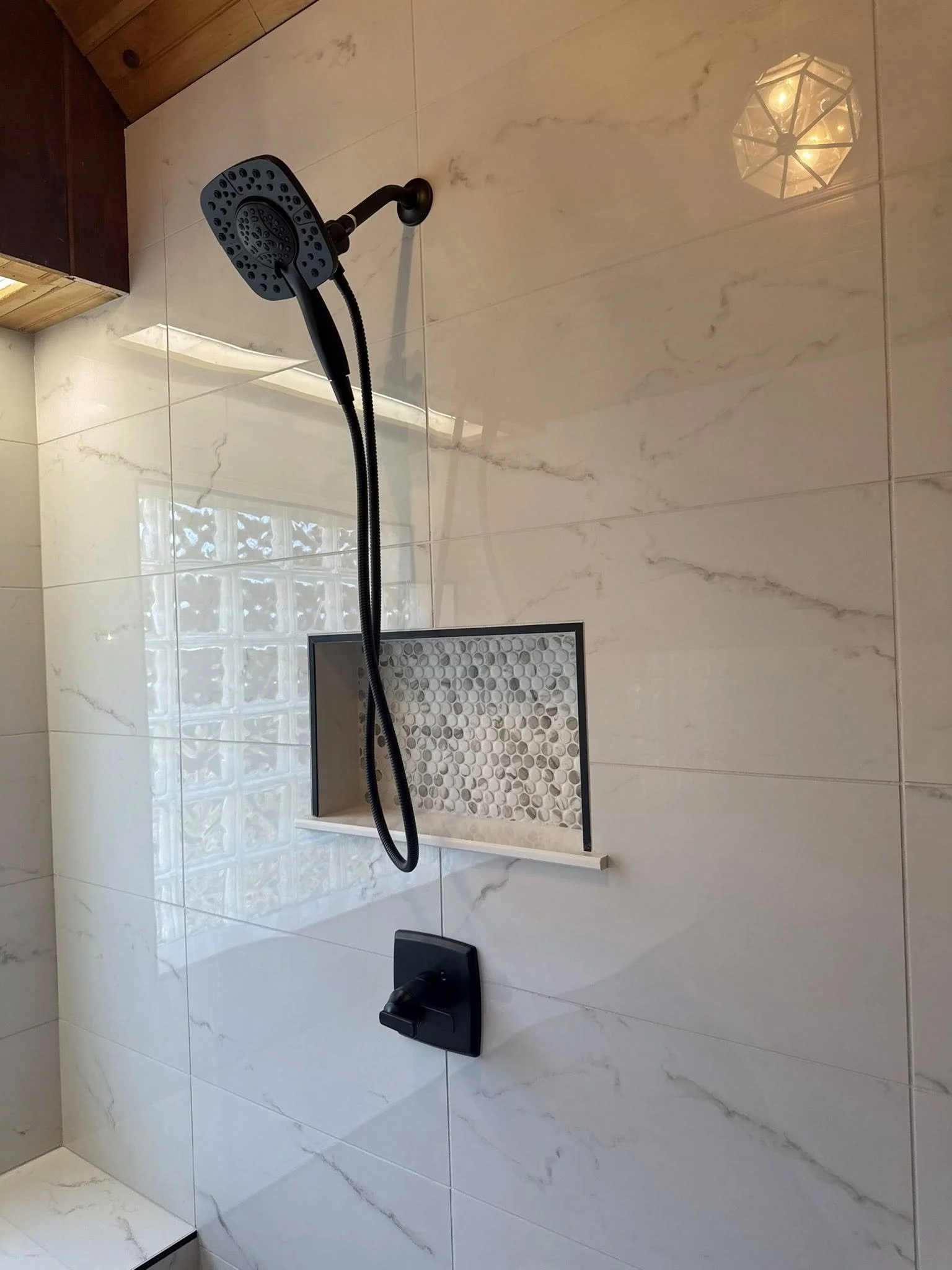 A modern walk-in shower with large white marble-like tiles, a round drain with pebble stones, a black showerhead attached to a black hose, and a geometric ceiling light fixture.