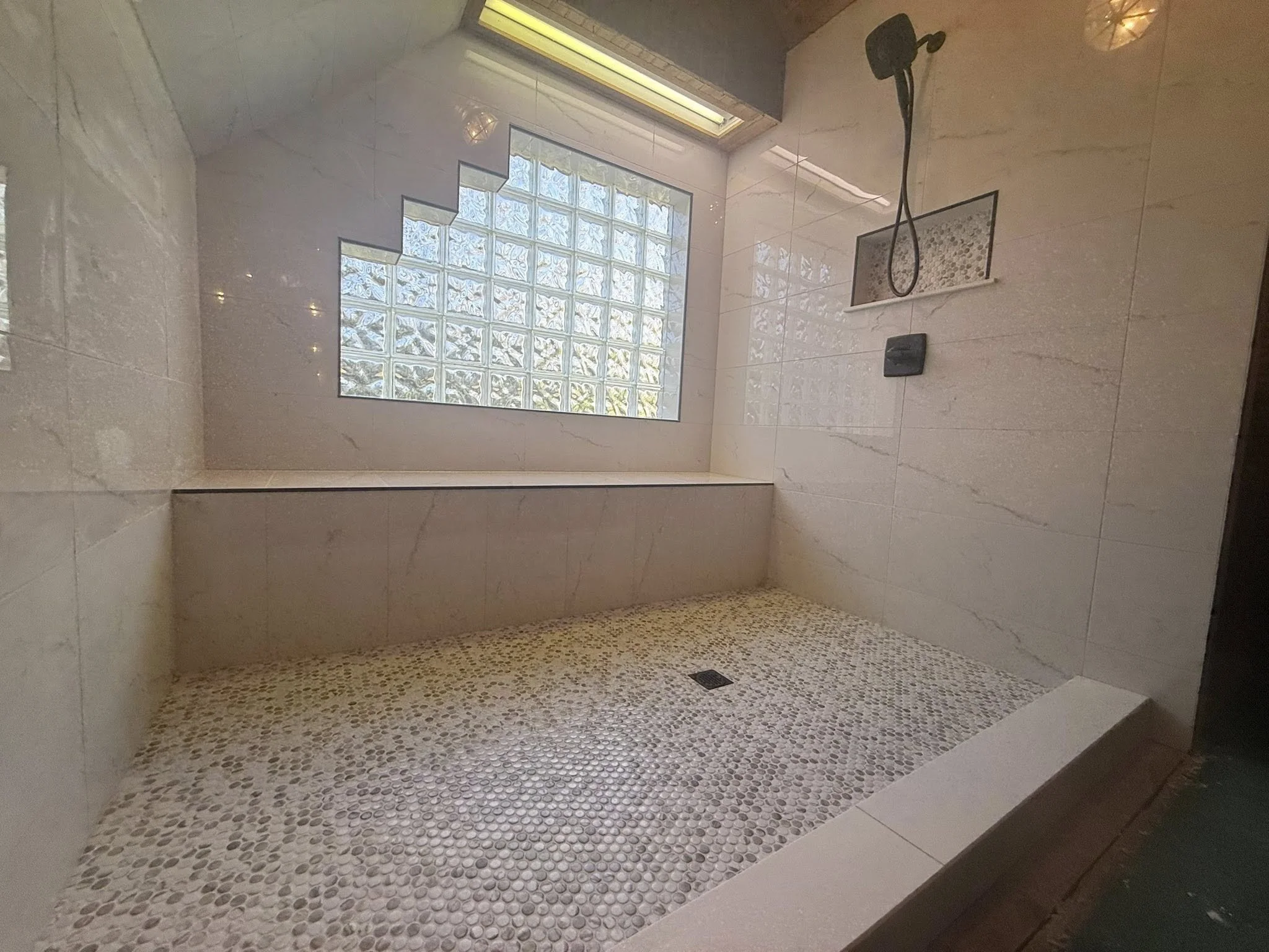 A modern walk-in shower with beige marble tile walls, a glass block window, and a pebble floor with a drain in the corner.