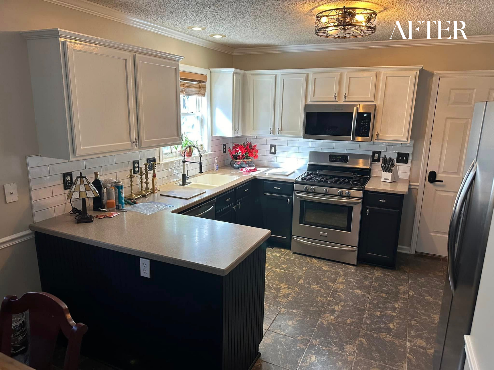 Kitchen after renovation with light upper cabinets, dark lower cabinets, stainless steel appliances, white subway tile backsplash, and a small window above the sink with a poinsettia plant. The ceiling has a decorative light fixture, and the island includes a small table lamp and various kitchen items.