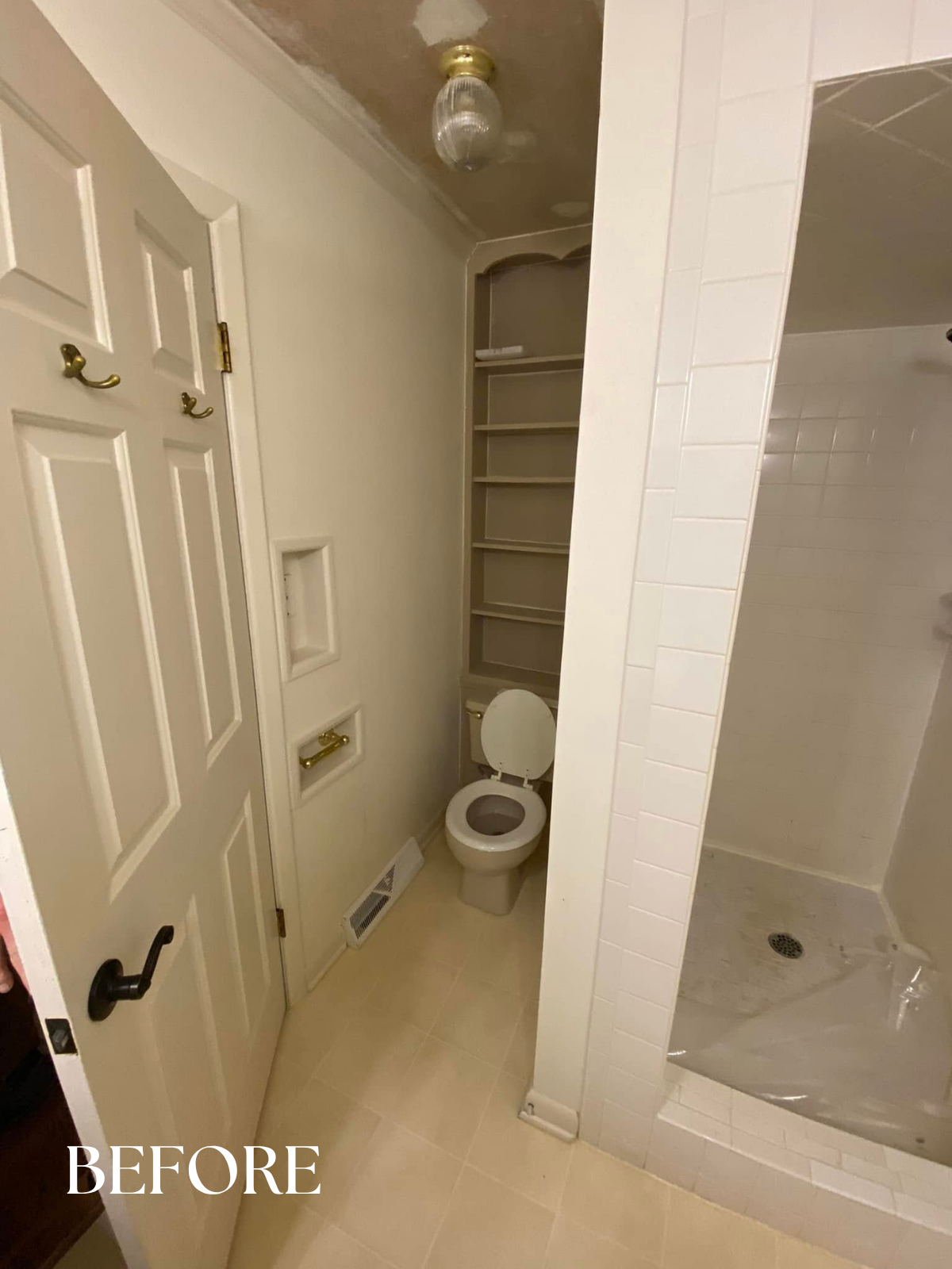 A small bathroom with a closed toilet, built-in shelves behind the toilet, a door with hooks, a vent on the floor, and a shower area with a drain, all in neutral tones.