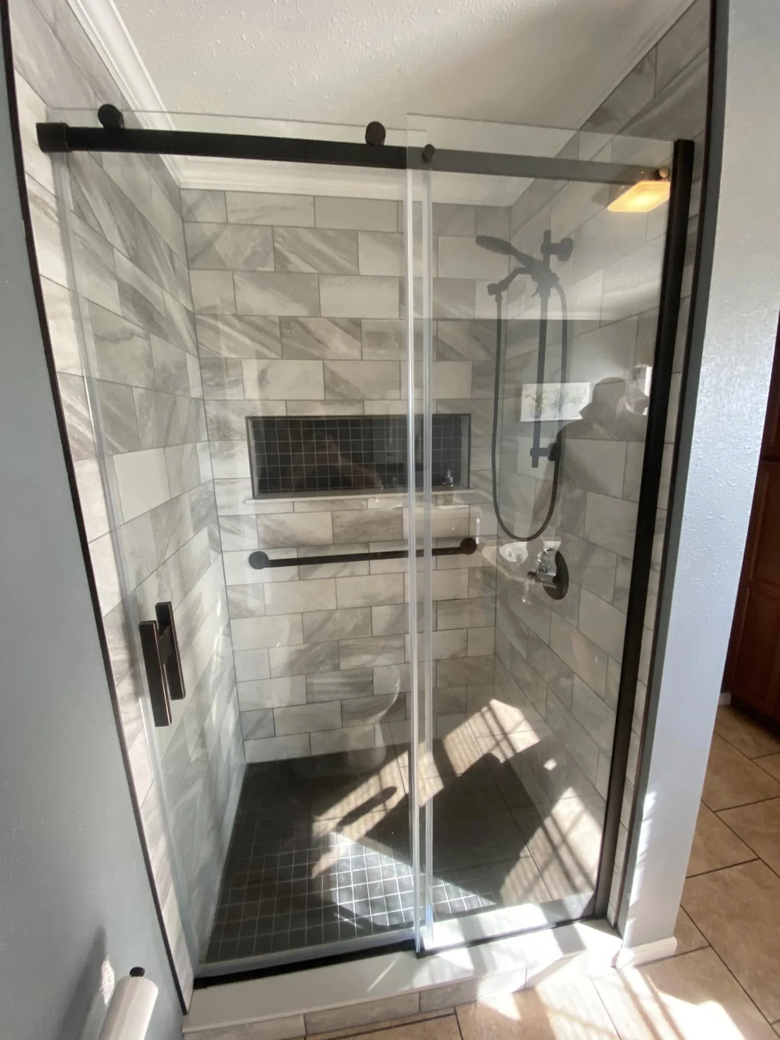 A modern walk-in shower with a glass door and black metal frame, tiled with beige and gray tiles, featuring a black handheld showerhead and a small window with black grid on the wall.