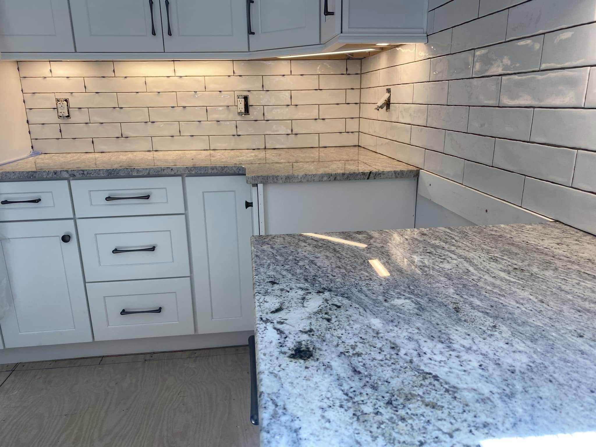 A kitchen with white cabinets, a gray granite countertop, a tile backsplash, and empty electrical outlets on the wall.