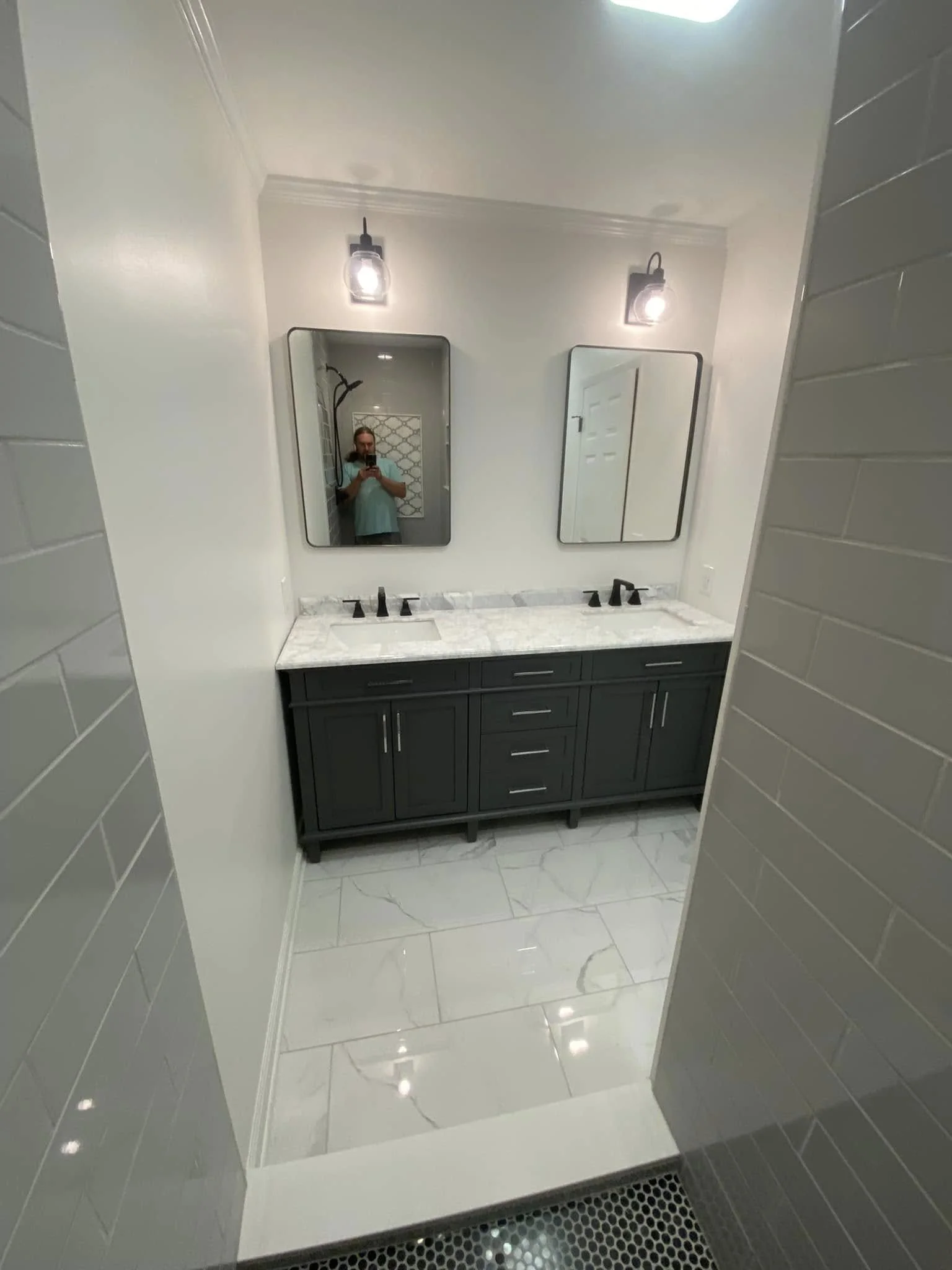 A modern bathroom vanity area with a dark gray cabinet, a white marble countertop, two black faucets, and two rectangular mirrors above each sink. Two wall-mounted light fixtures are above the mirrors, and the bathroom has white tile walls and floori