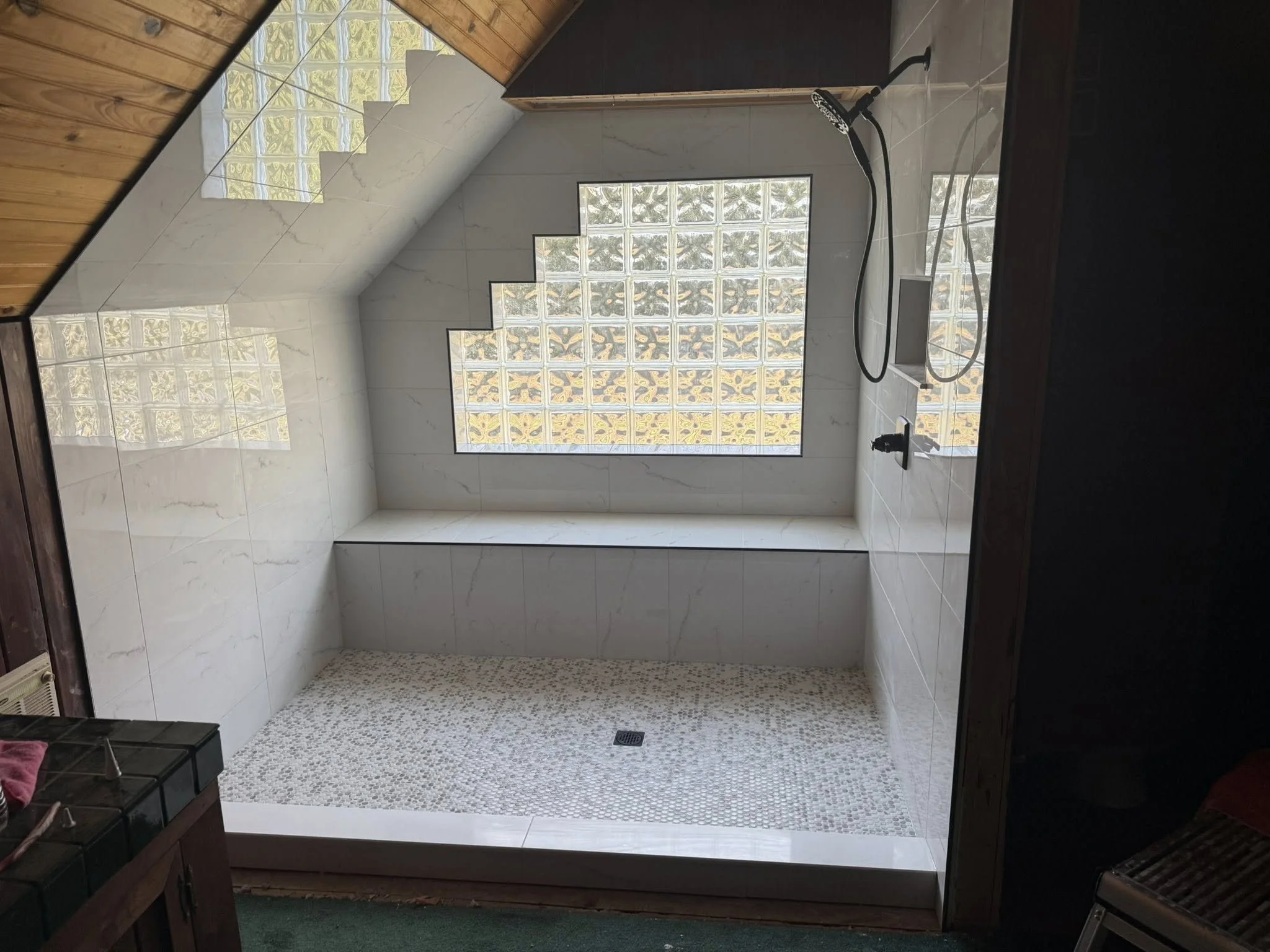 View of a walk-in shower with a glass block wall, white marble tile walls, a wooden ceiling, a shower drain on the floor, a small window, and part of a black shower fixture visible.