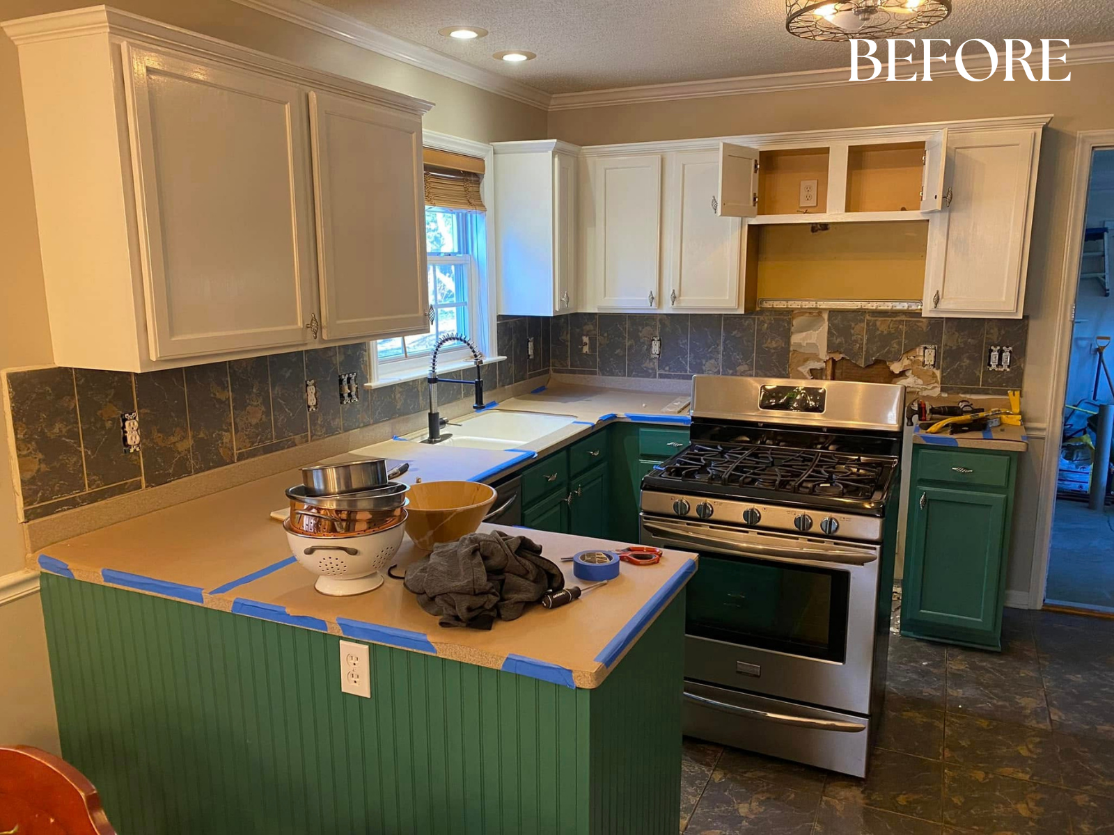 Kitchen with white upper cabinets and green lower cabinets, a stainless steel stove, a black countertop, a window above the sink, and various tools and items on the countertop, indicating ongoing renovation.