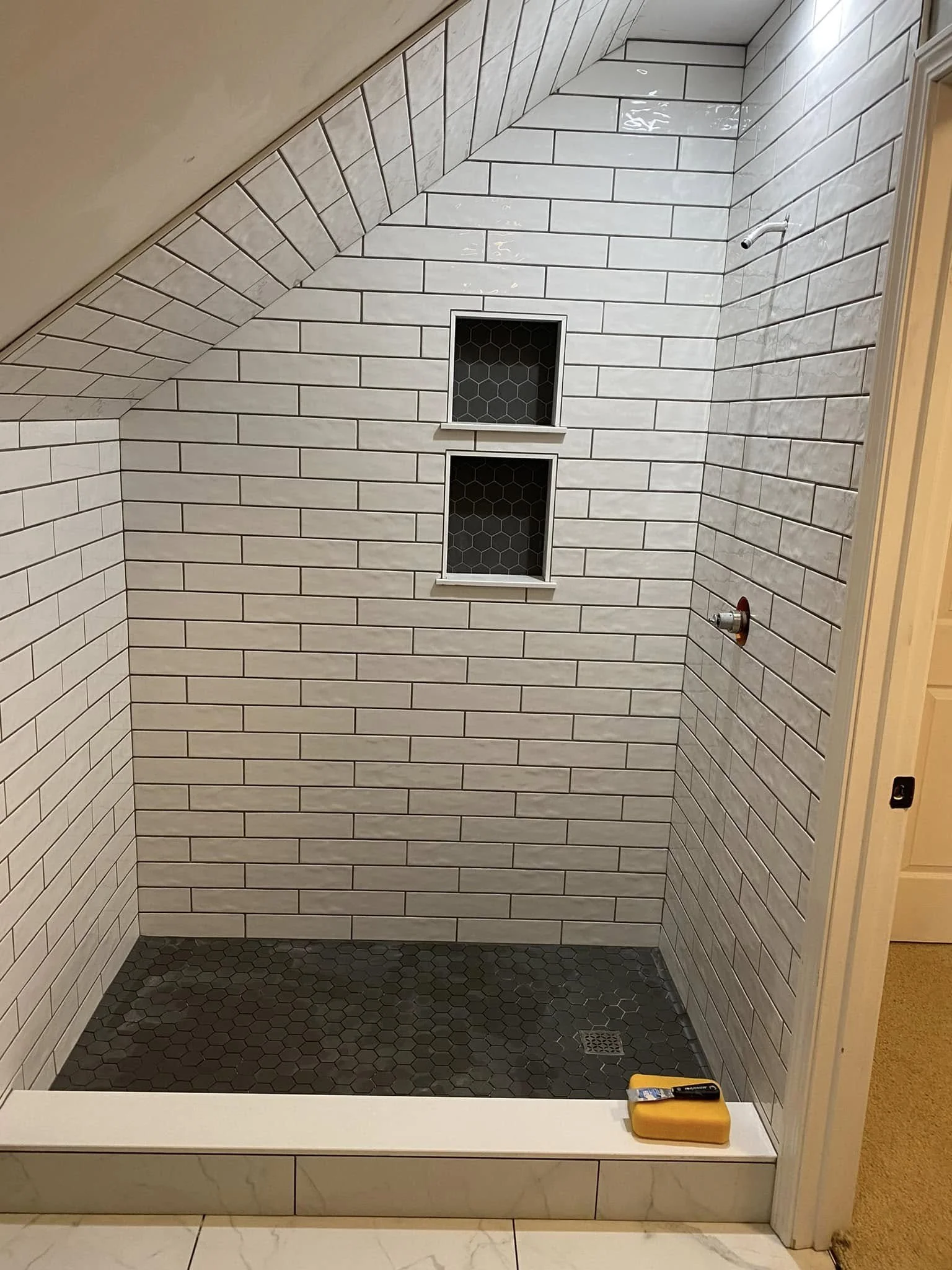 An empty walk-in shower with white subway tile walls, a small built-in shelf with black hex tile background, a small window, a pipe for a showerhead, and black hex tiles on the floor.