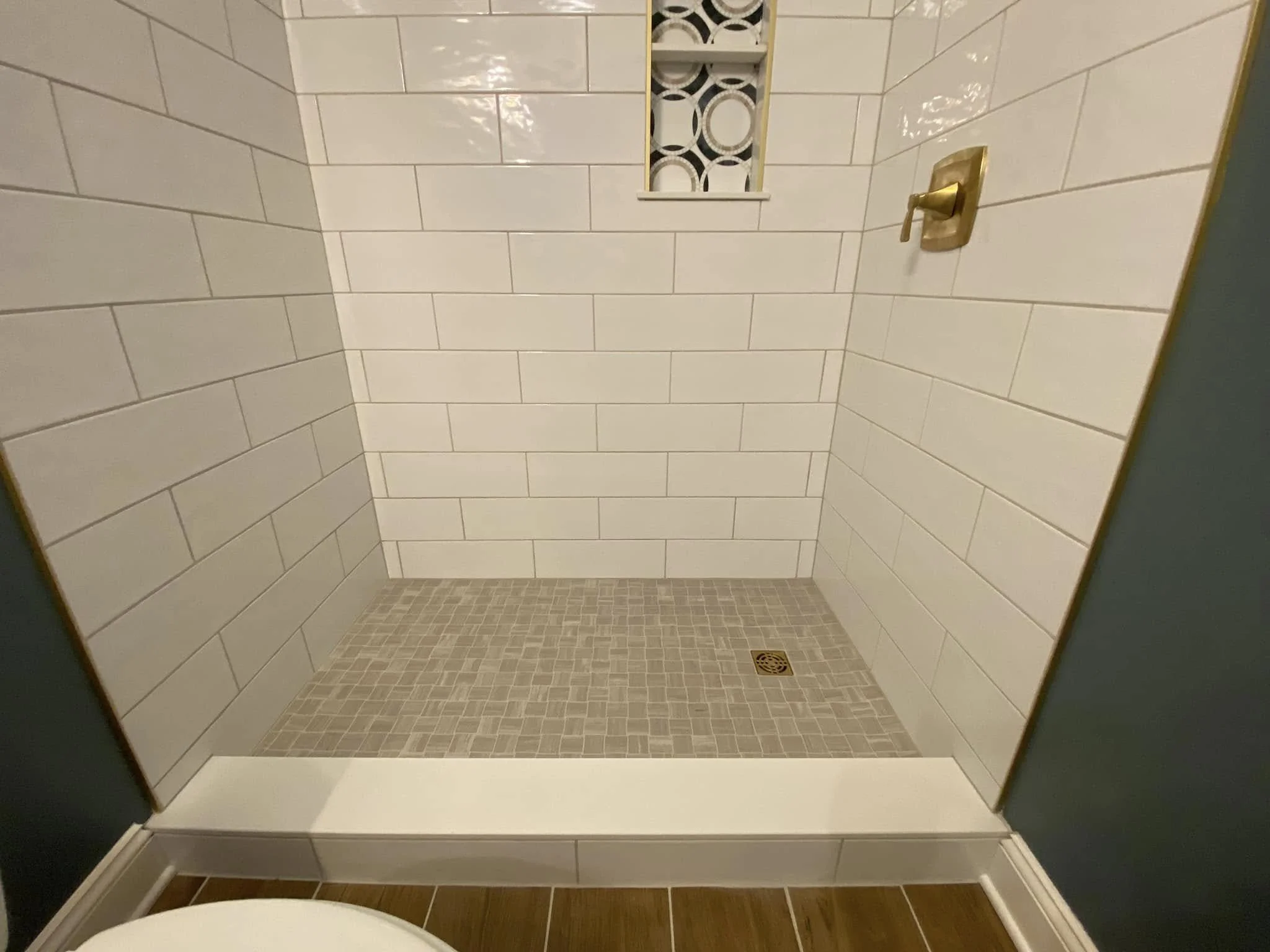 A shower stall with white subway tiles on the walls, small beige mosaic tiles on the floor, a brass shower handle, a small window with black and white geometric patterned glass, and a floor drain.