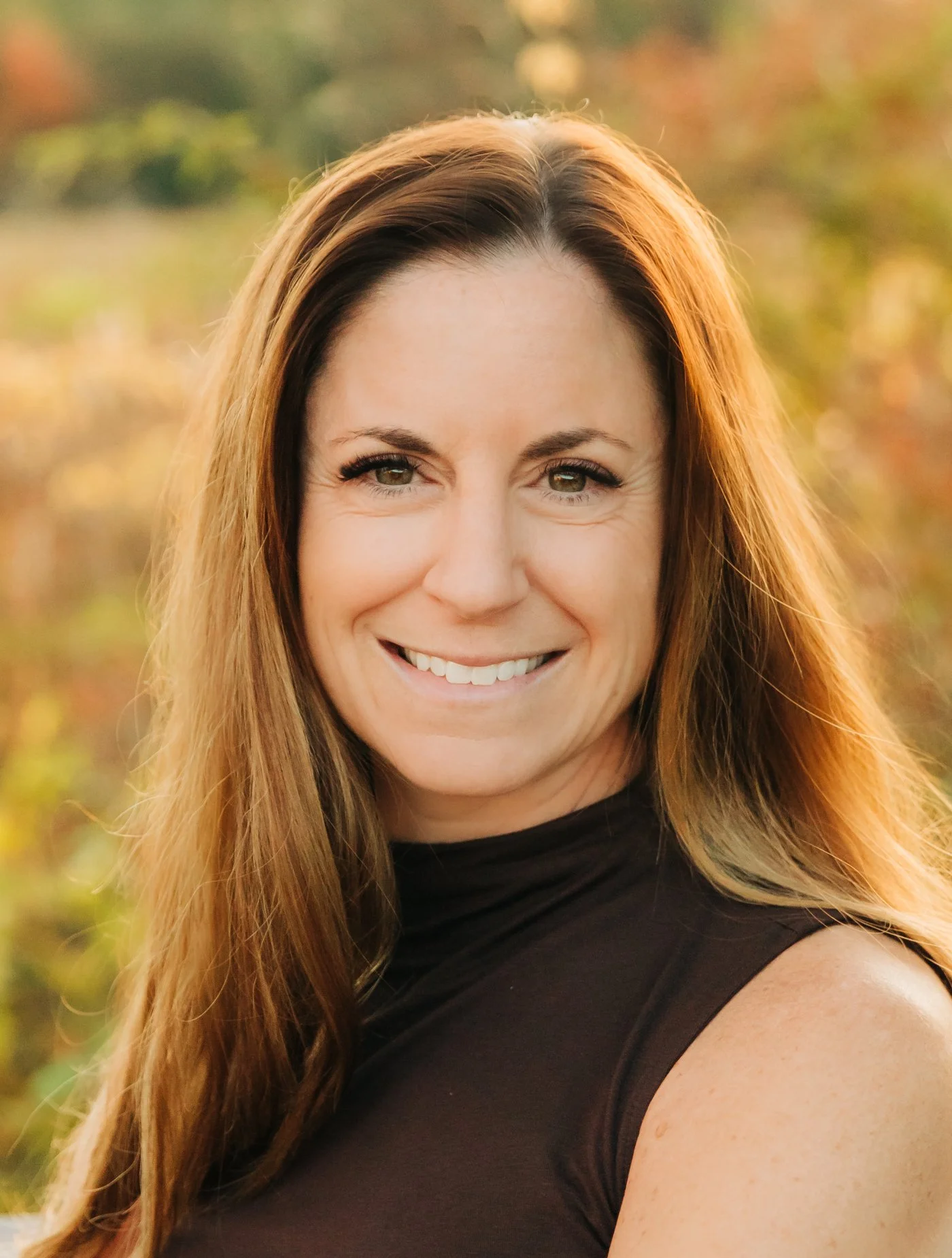 A woman with long, reddish-brown hair and green eyes smiling outdoors with a colorful, blurred autumn background.