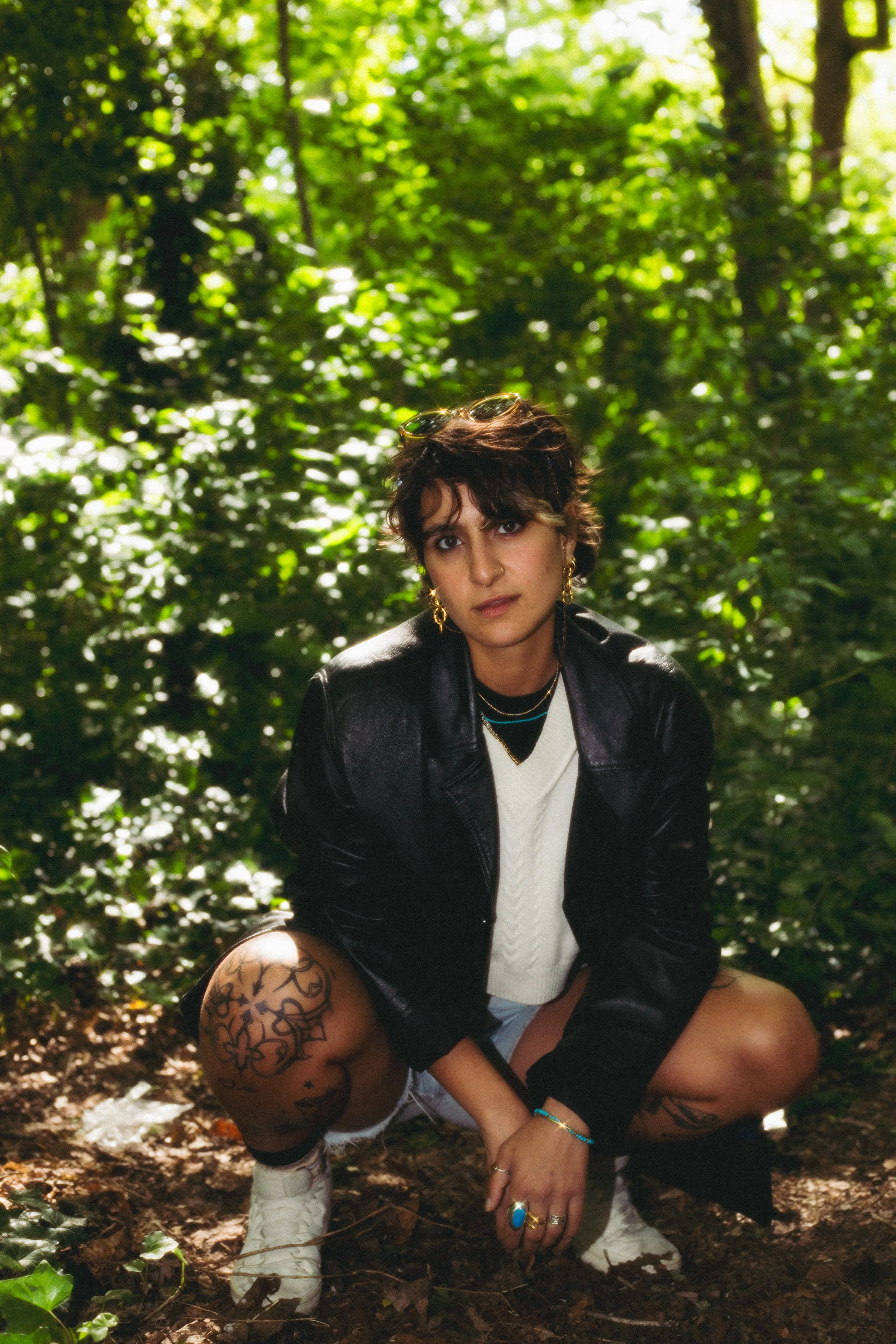 A young woman with short dark hair, tattoos on her left thigh, multiple rings and earrings, and sunglasses on her head, squatting in a forest with green foliage and sunlight filtering through the trees, wearing a black leather jacket, a white sweater, and shorts.