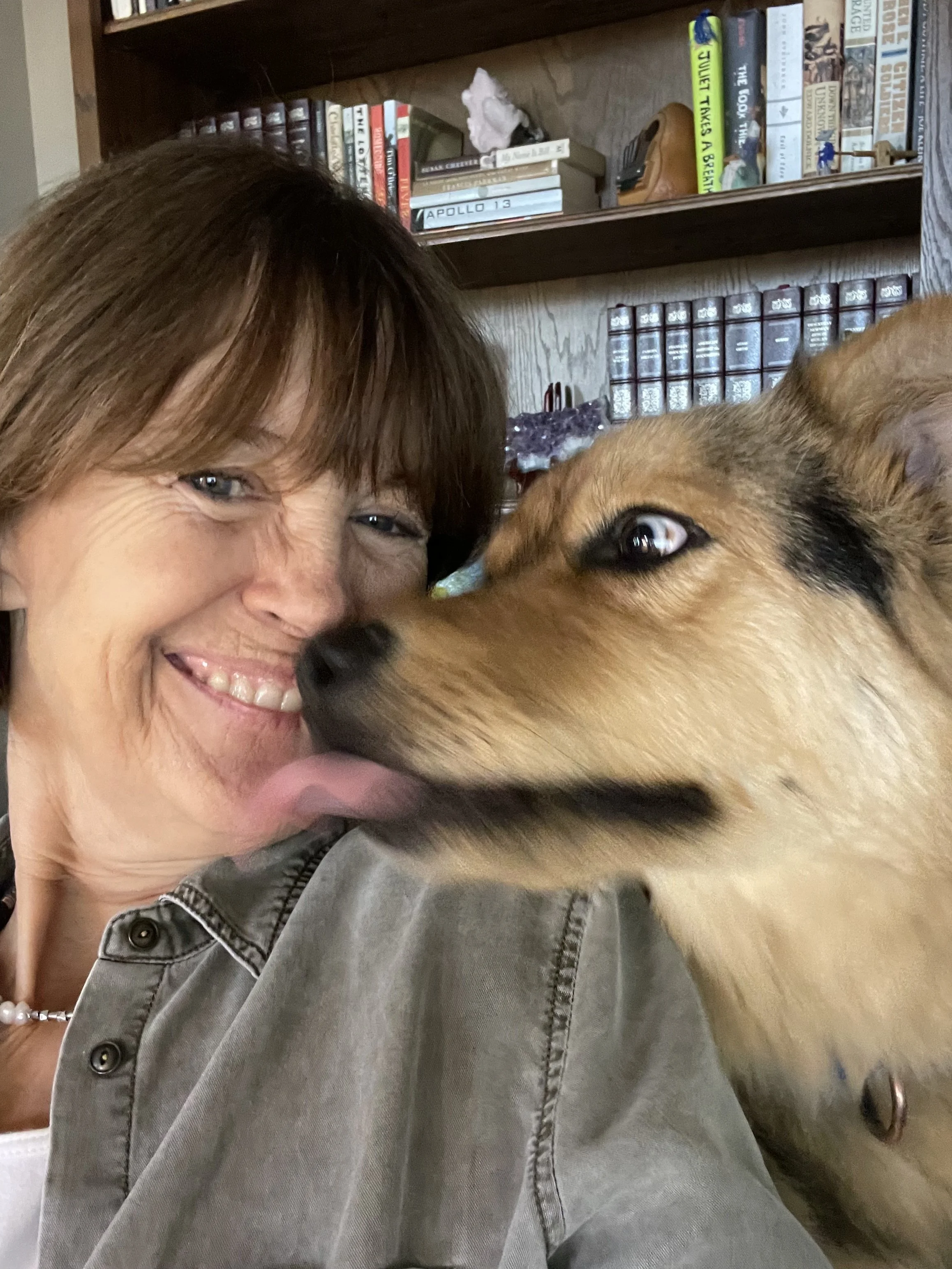A woman with short brown hair and a gray jacket smiling with a dog that has tan fur and black markings, in a room with wooden bookshelves filled with books.